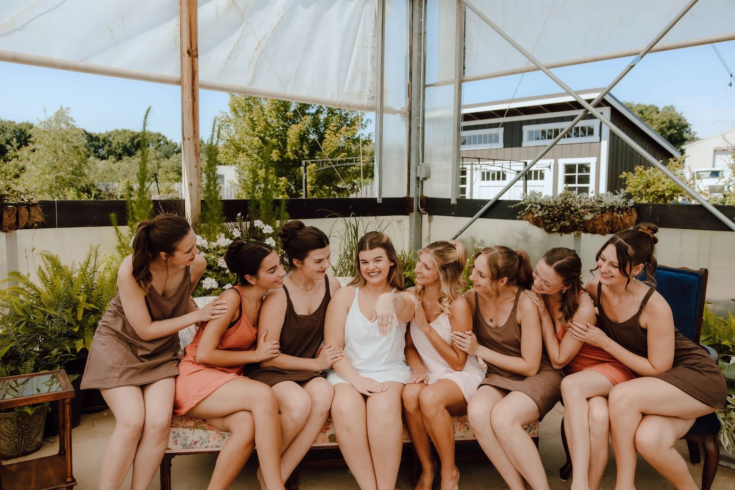 Bride in white surrounded by bridesmaids in earthy tones sitting together laughing outdoors at a greenhouse venue.