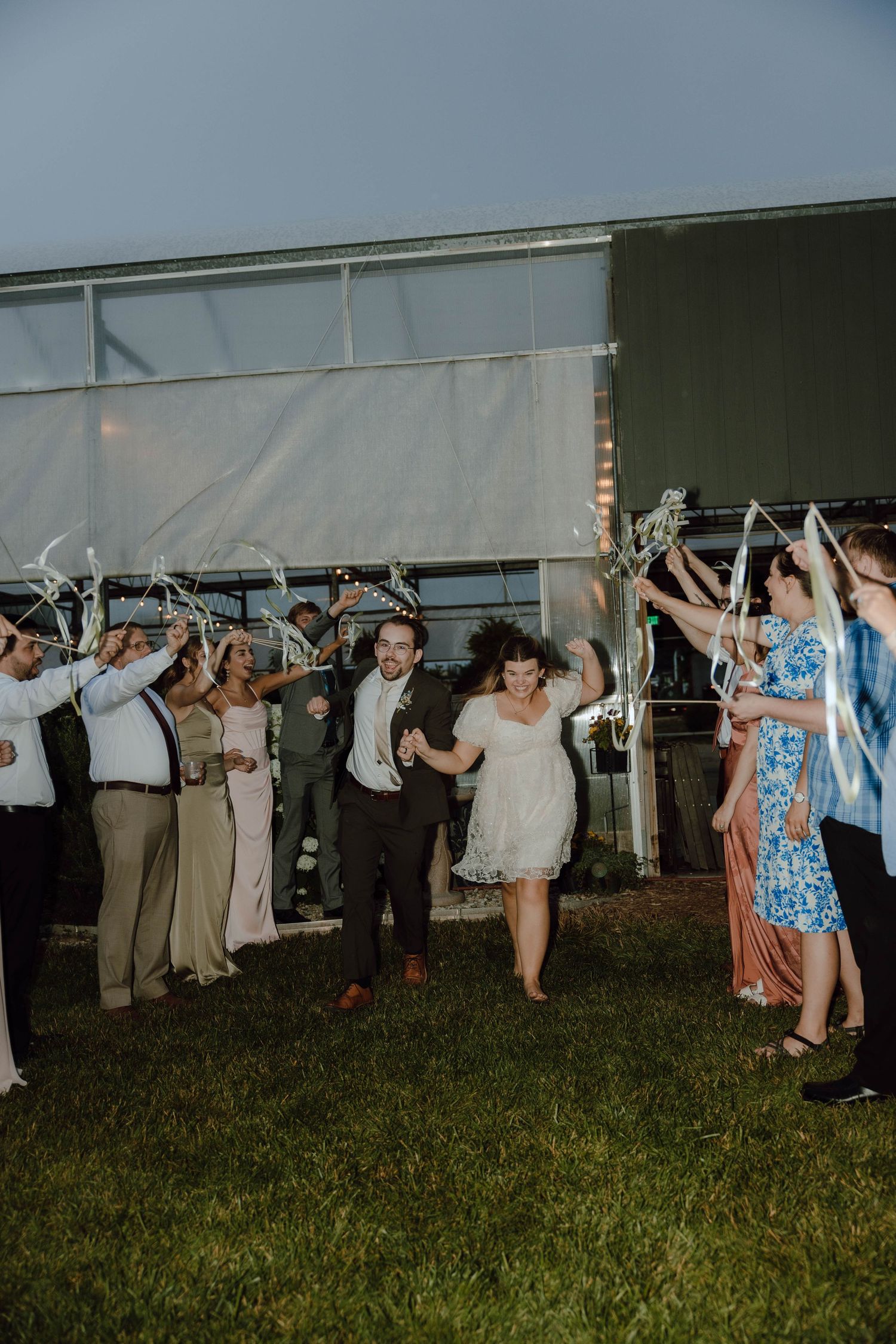 Bride and groom exit wedding reception through ribbon wand send-off tunnel with cheering guests outdoors at dusk.