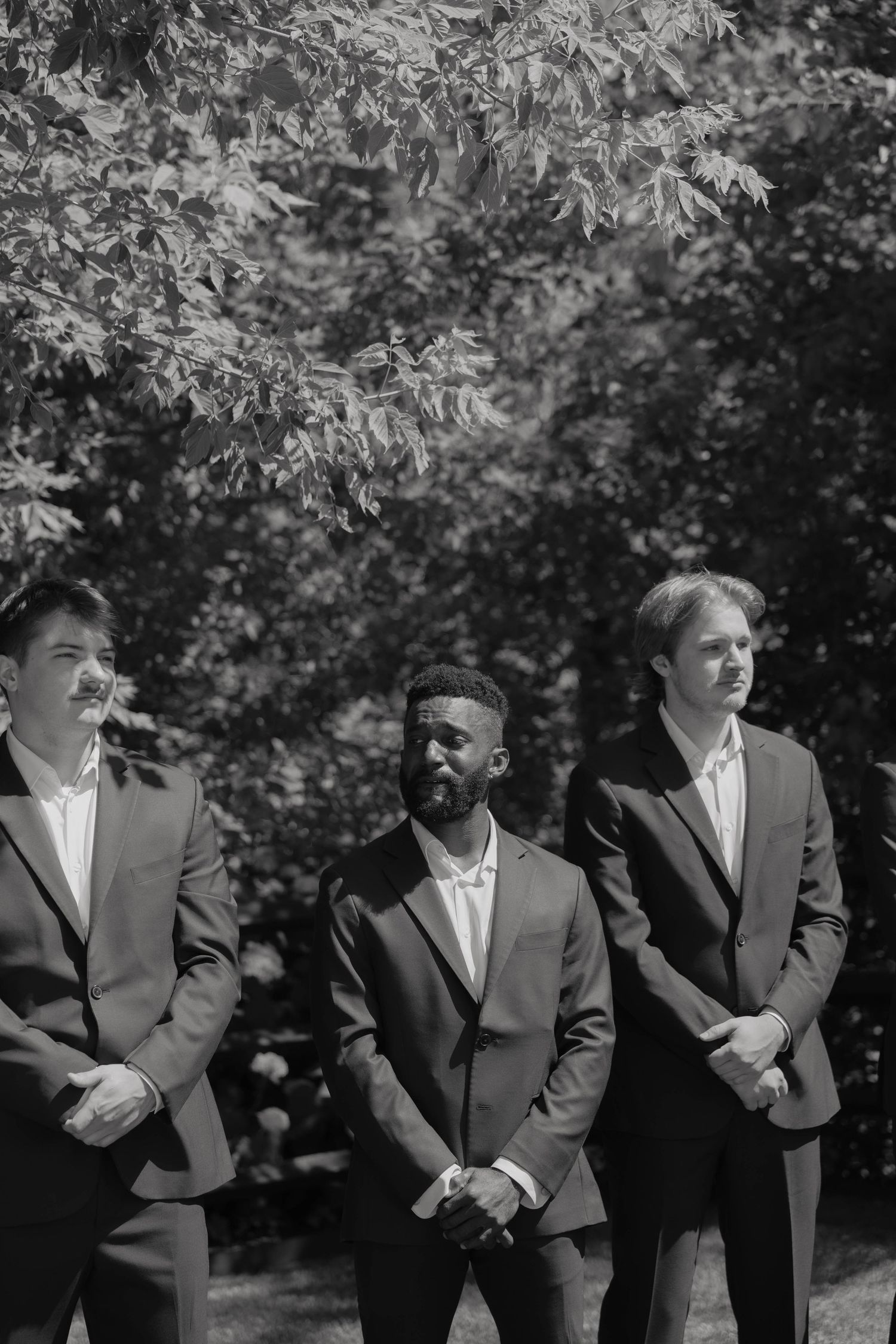 Three groomsmen in dark suits stand outdoors among lush trees in a black and white wedding photo.