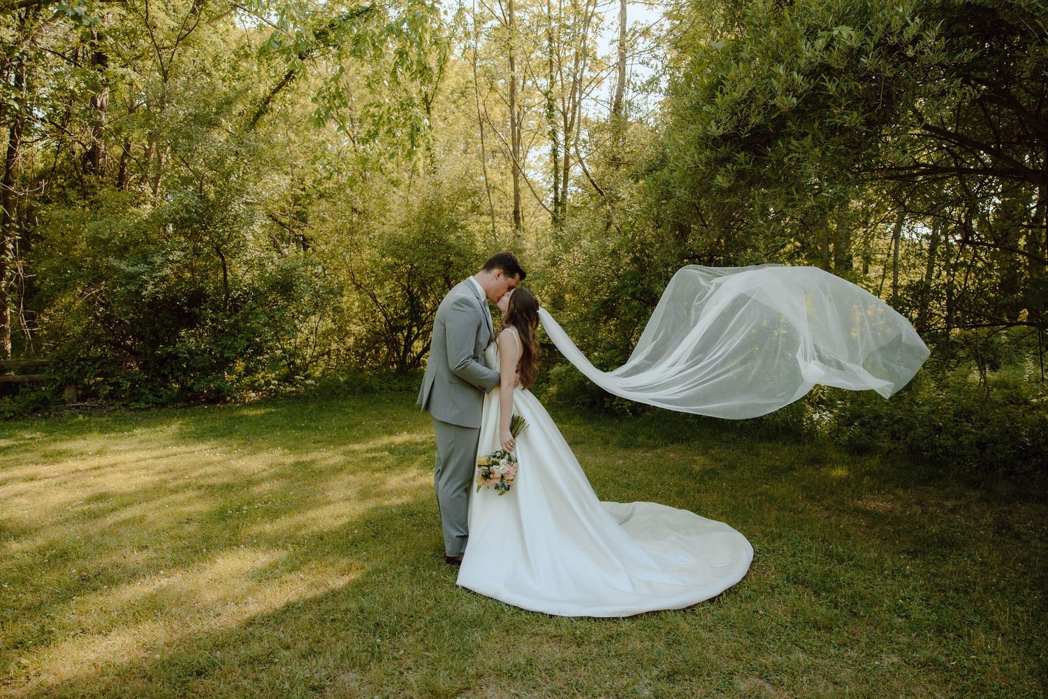 Bride and groom share a romantic kiss outdoors as her veil flows gracefully in the wind amid lush green trees.