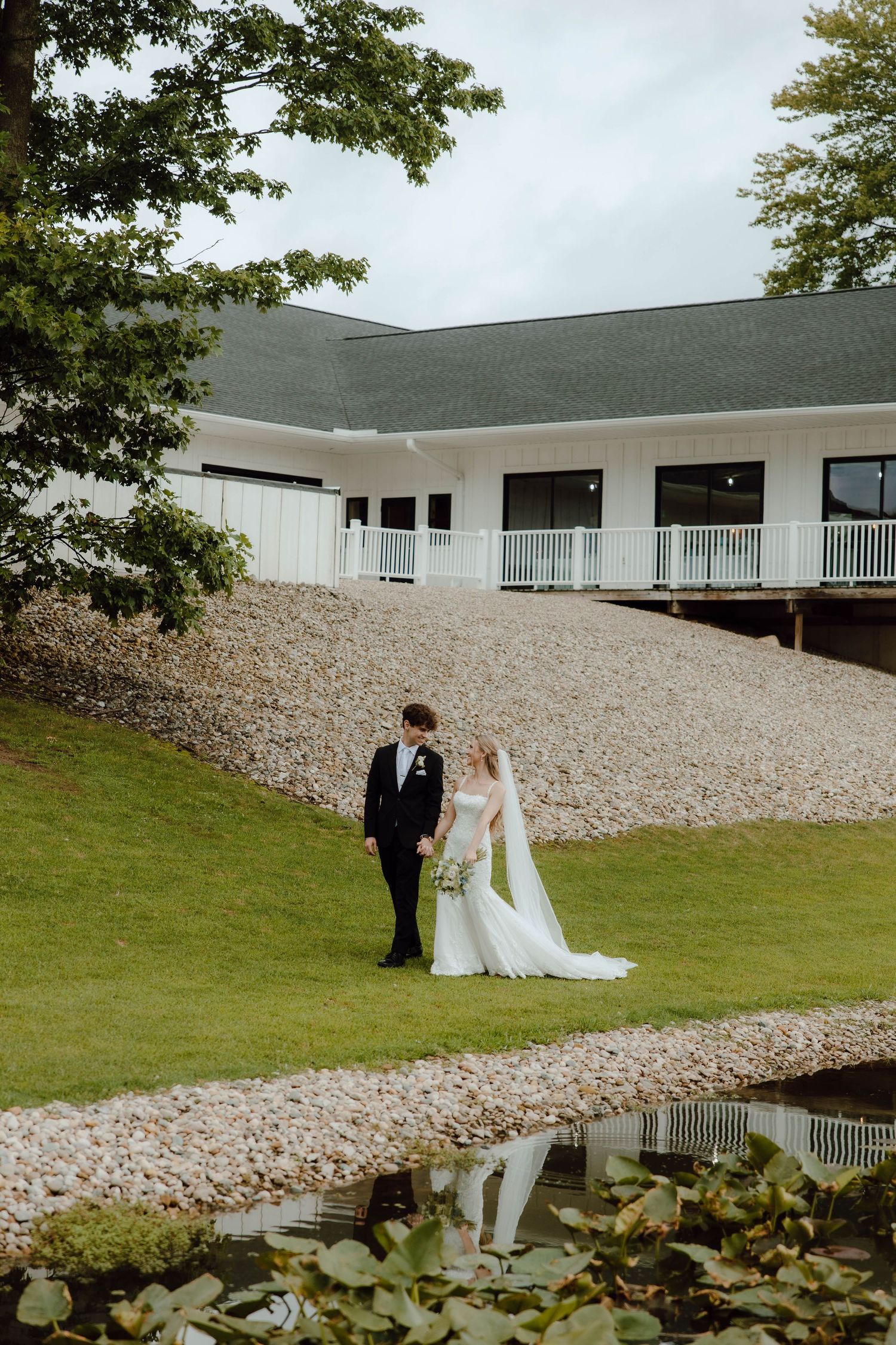 Bride and groom stand by a serene pond on lush green grounds outside an elegant white wedding venue.