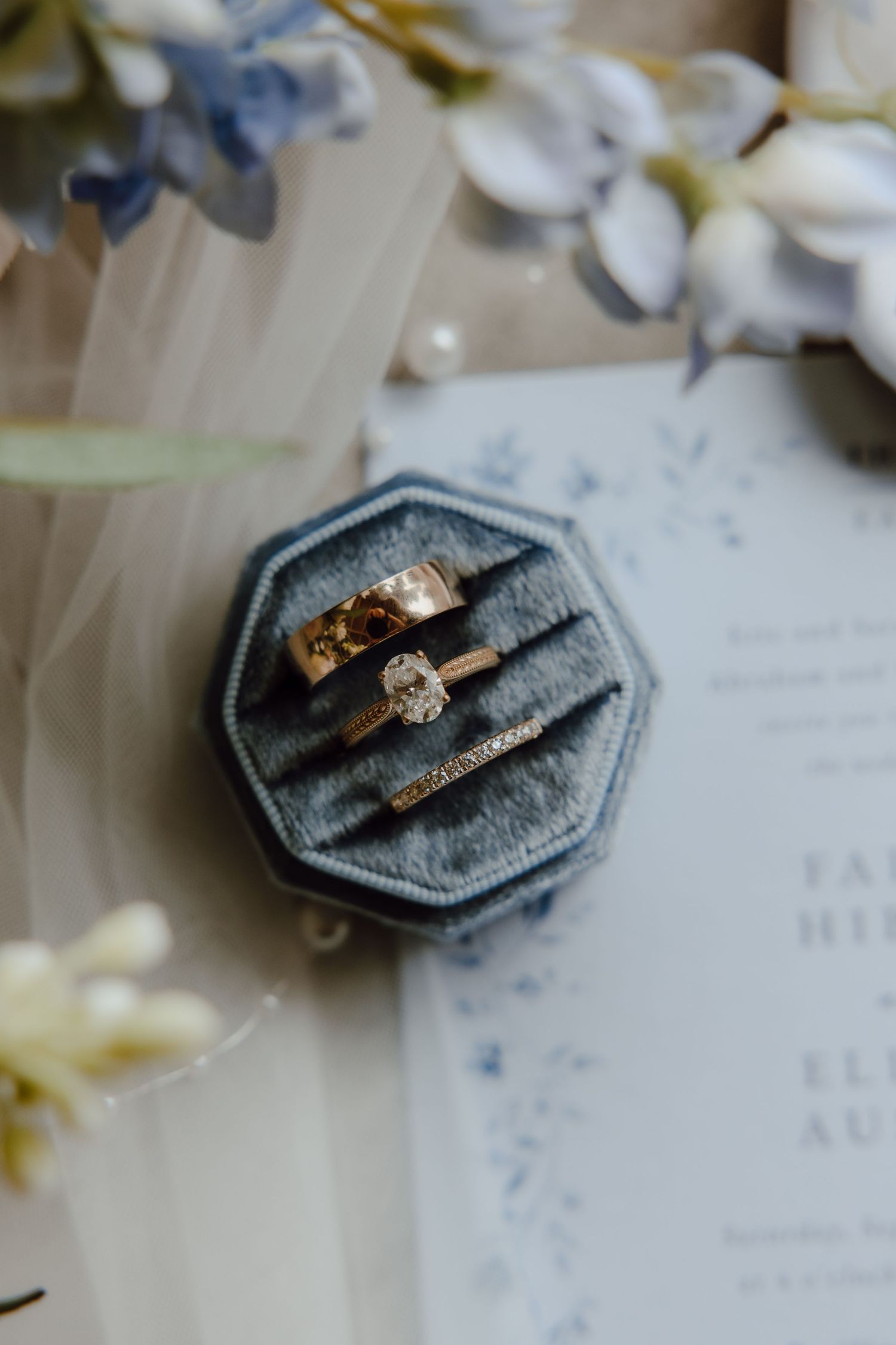 Gold wedding rings displayed in a velvet ring box with blue florals and wedding invitation.