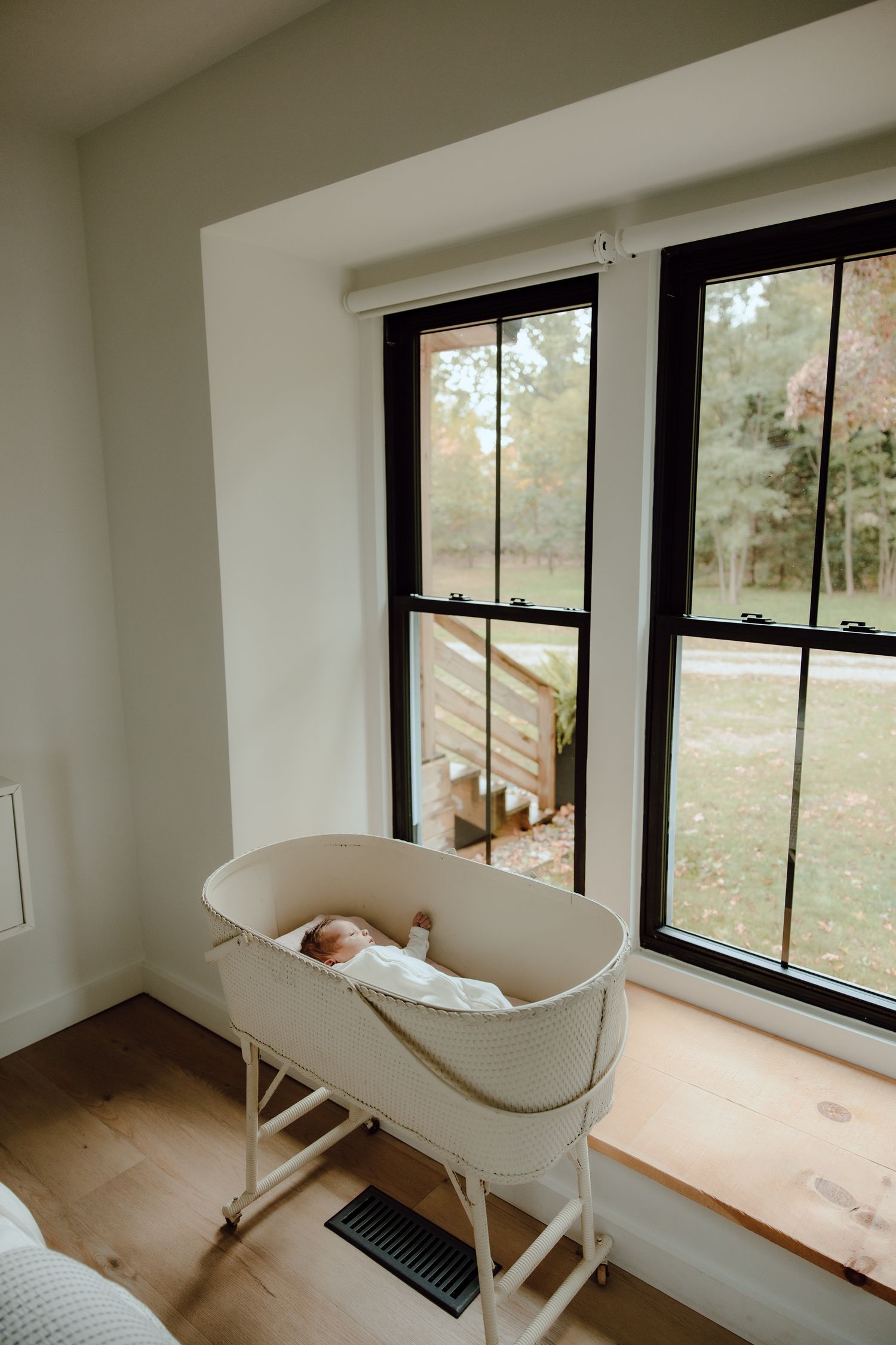 White bassinet by large windows in a minimalist nursery with natural light and wooden floors.