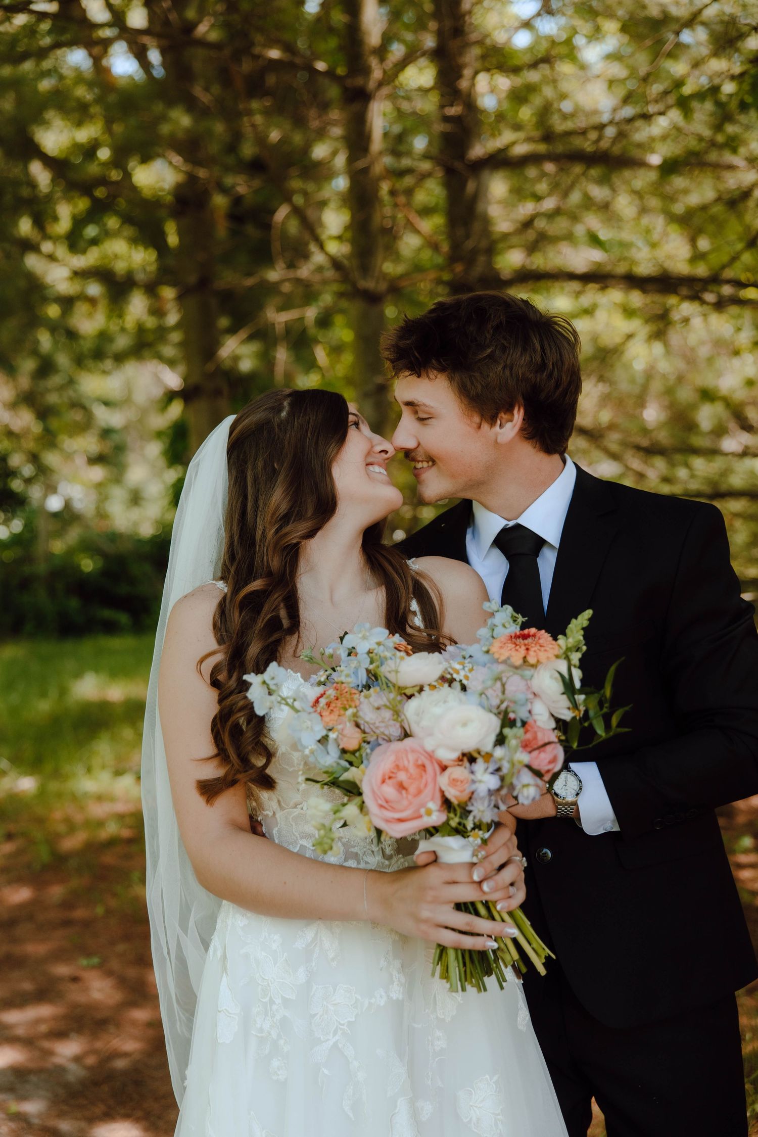 A bride and groom share a romantic moment outdoors, smiling at each other while holding a pastel floral bouquet.