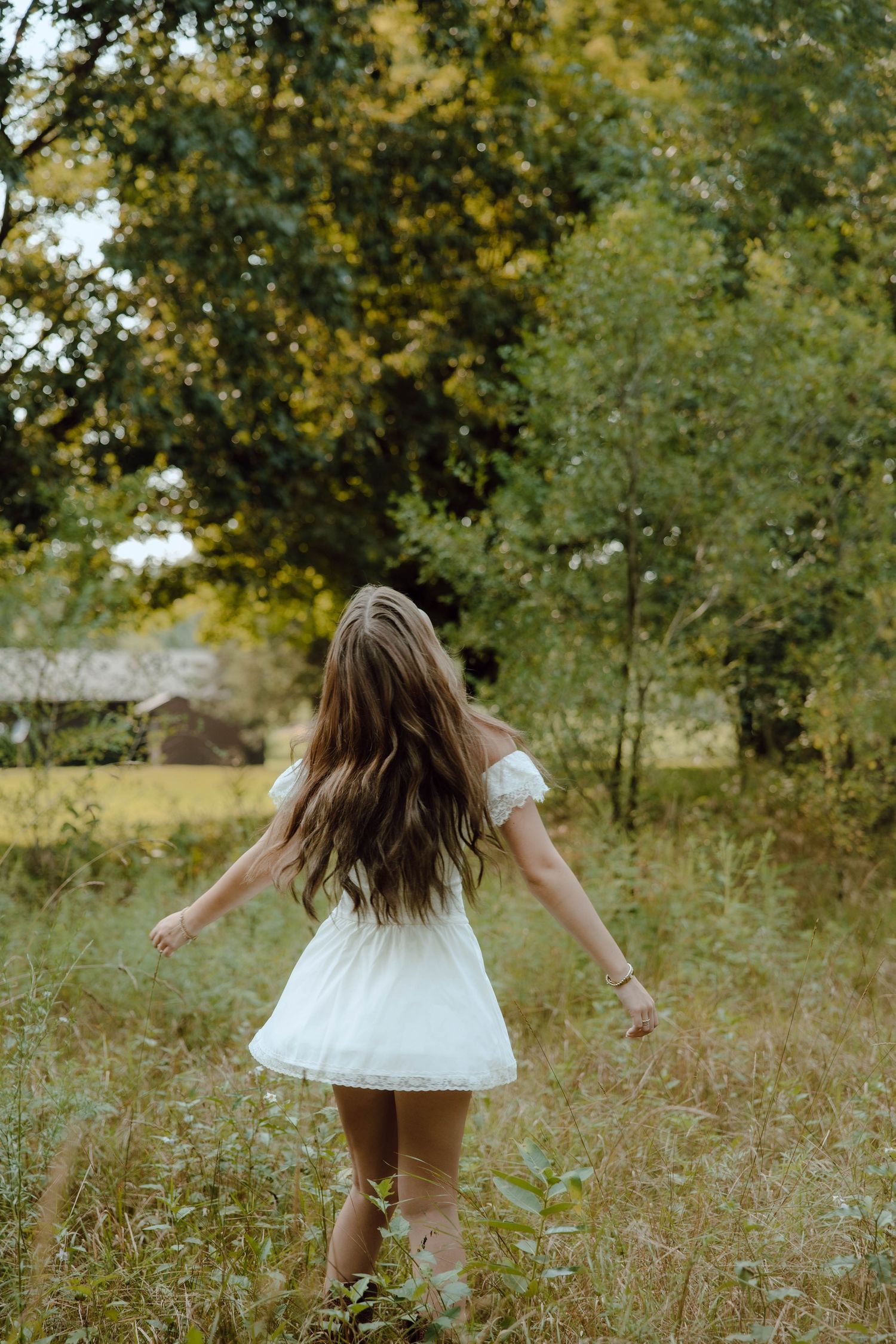 Young woman in white mini dress walks through lush green meadow toward trees, long brown hair flowing freely.