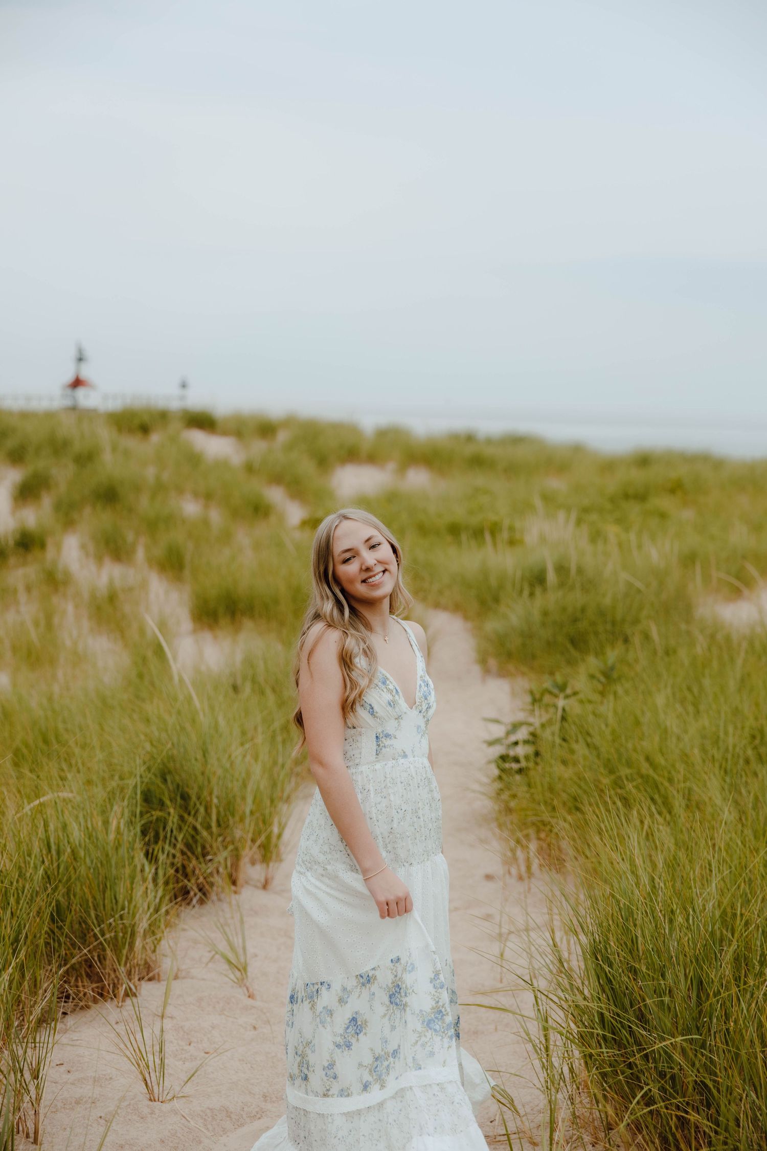 Smiling blonde woman in a floral maxi dress standing on a sandy beach path surrounded by tall green grass.