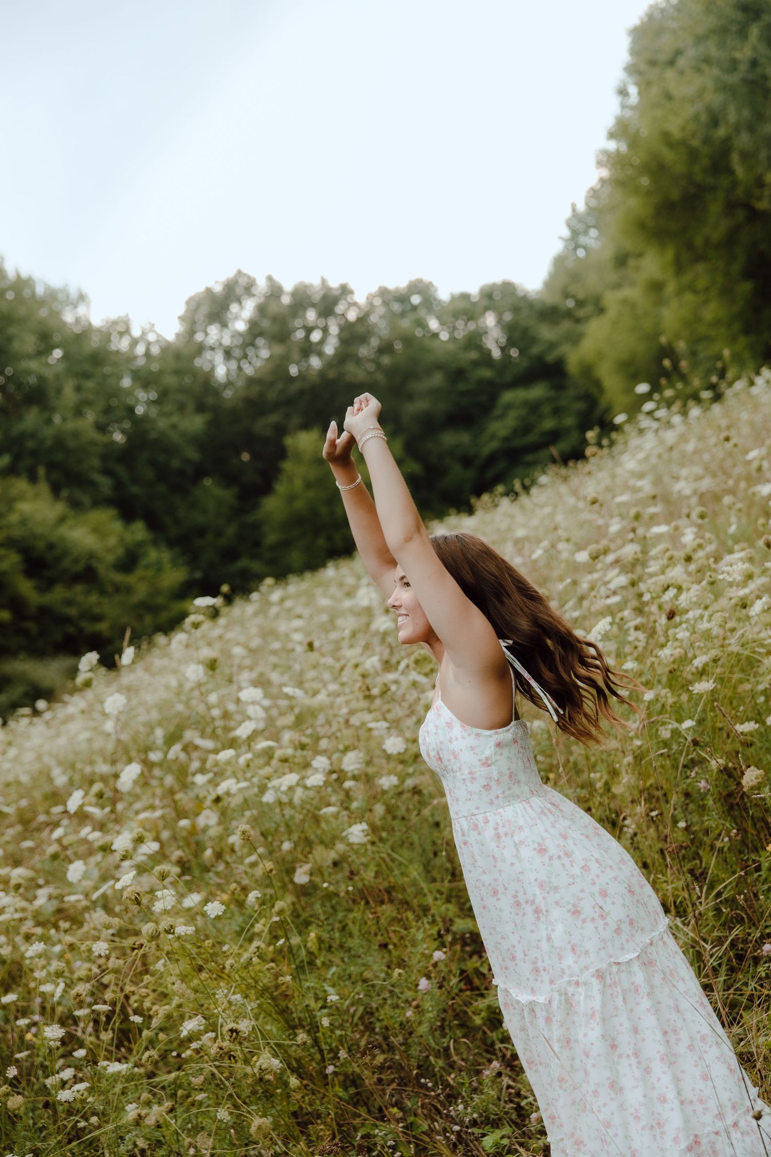 Young woman in white floral dress raising arm joyfully in a wildflower meadow with lush green trees.