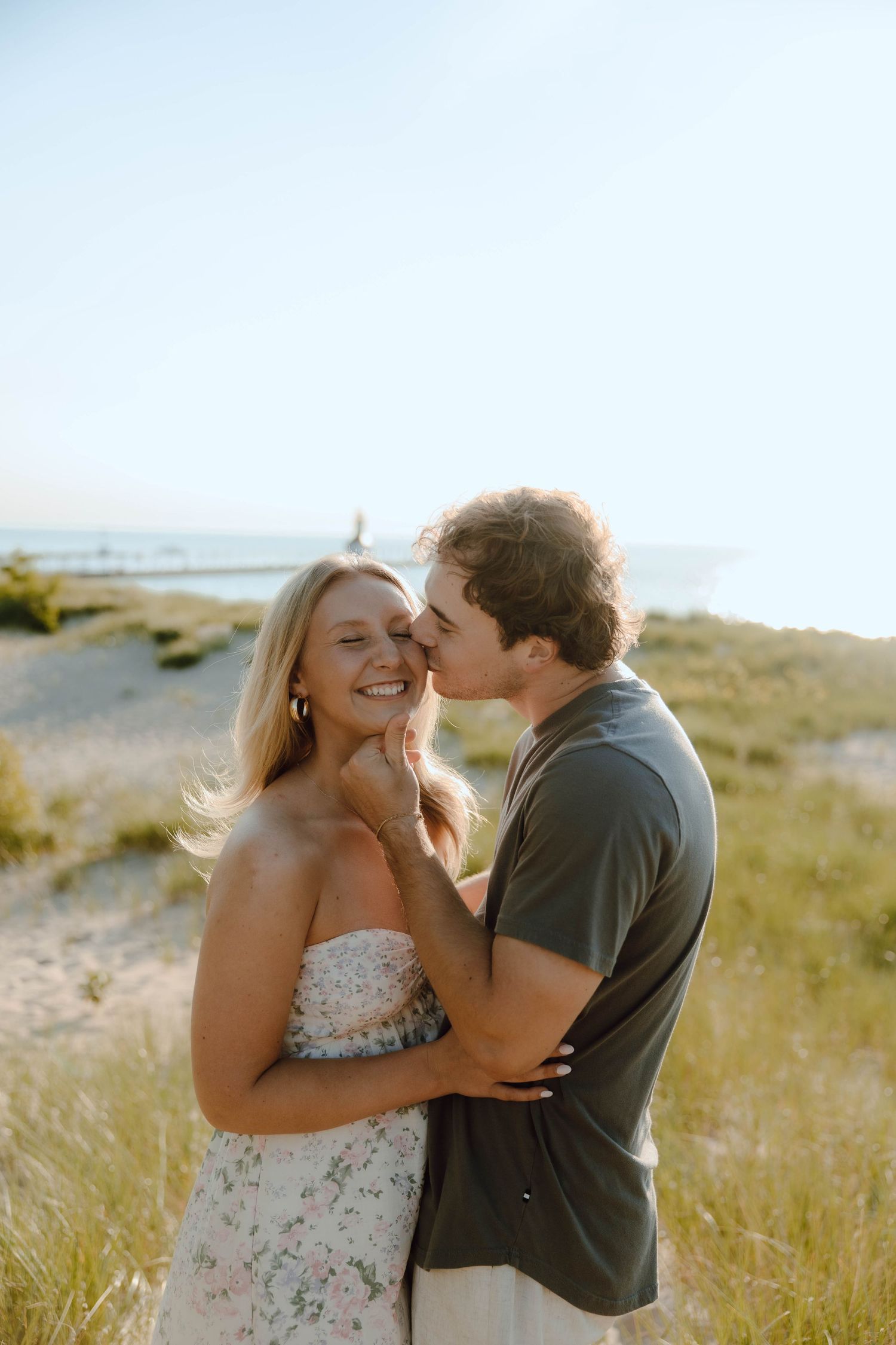 A couple shares a tender kiss on a sunny beach, with the man gently holding the smiling woman's face near coastal dunes.