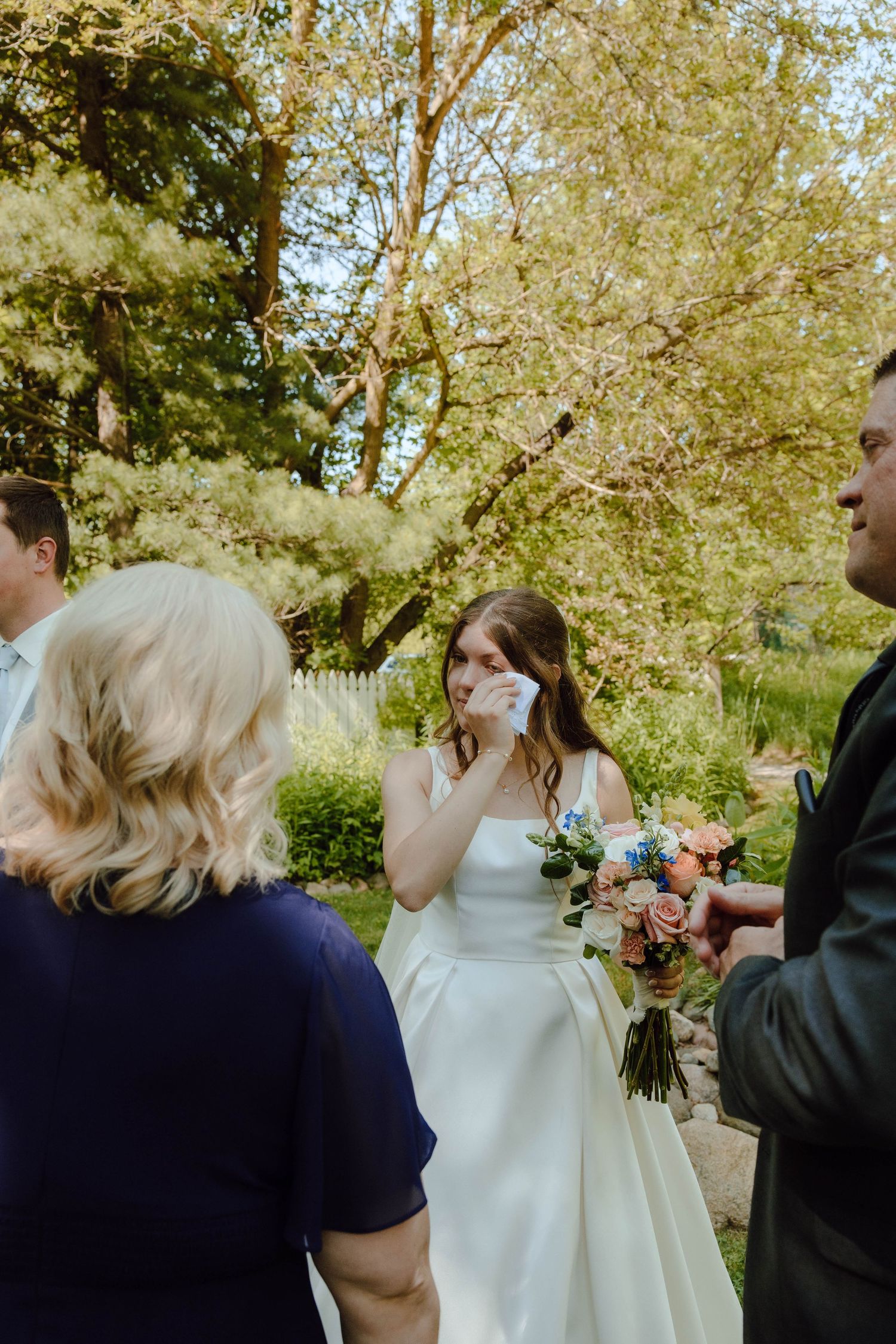 Emotional bride wipes tears during outdoor wedding ceremony while holding a pastel pink and blue bouquet.