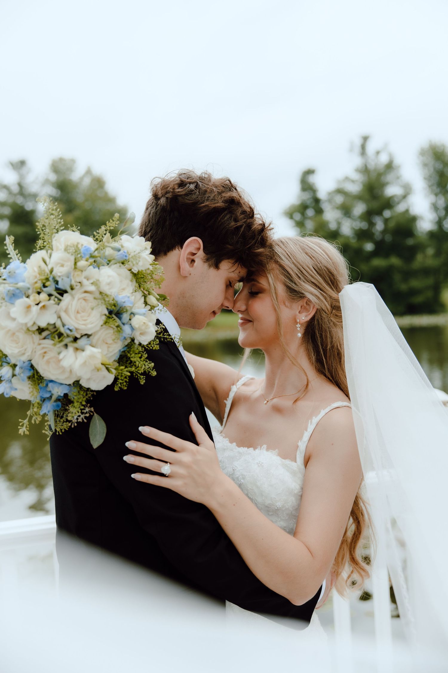 A bride and groom share a romantic moment, nearly kissing, as she holds a blue and white bouquet outdoors.
