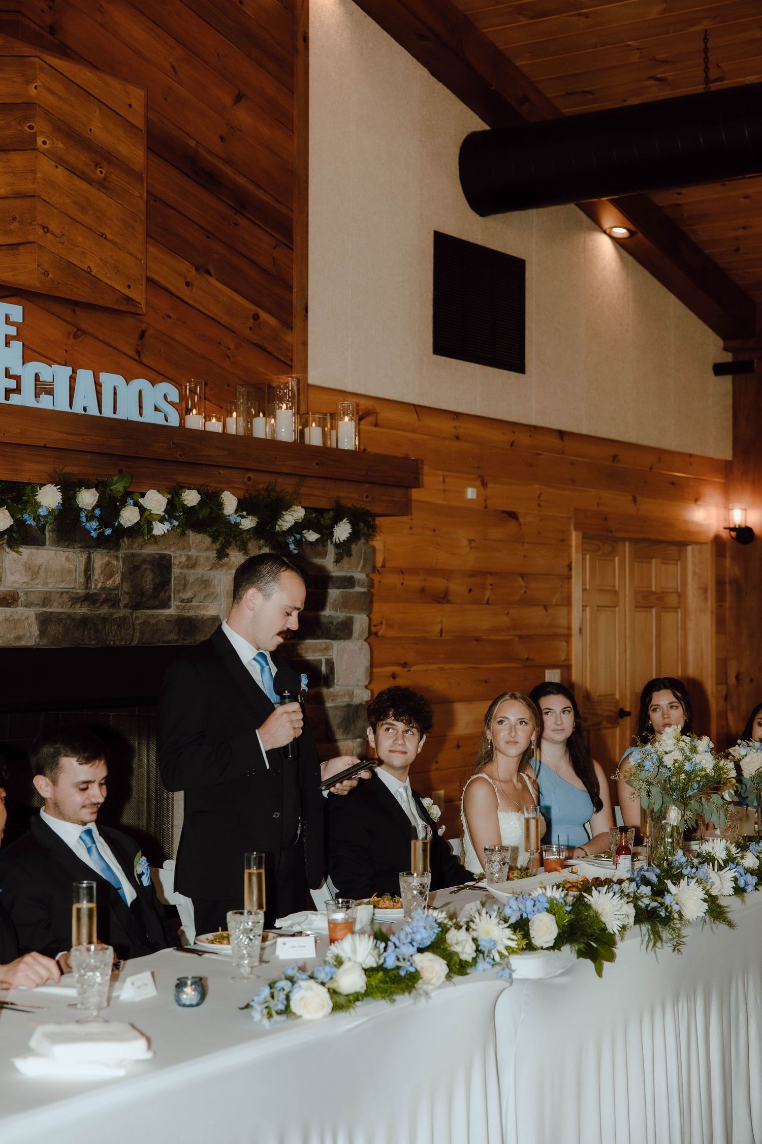 Man giving speech at wedding reception in rustic wood-paneled venue with floral arrangements.