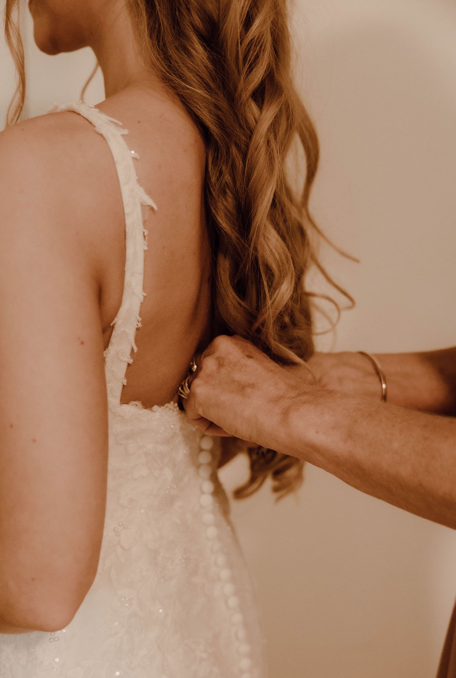 A bride in a white lace wedding dress has the back fastened by a helper with a gold bracelet.