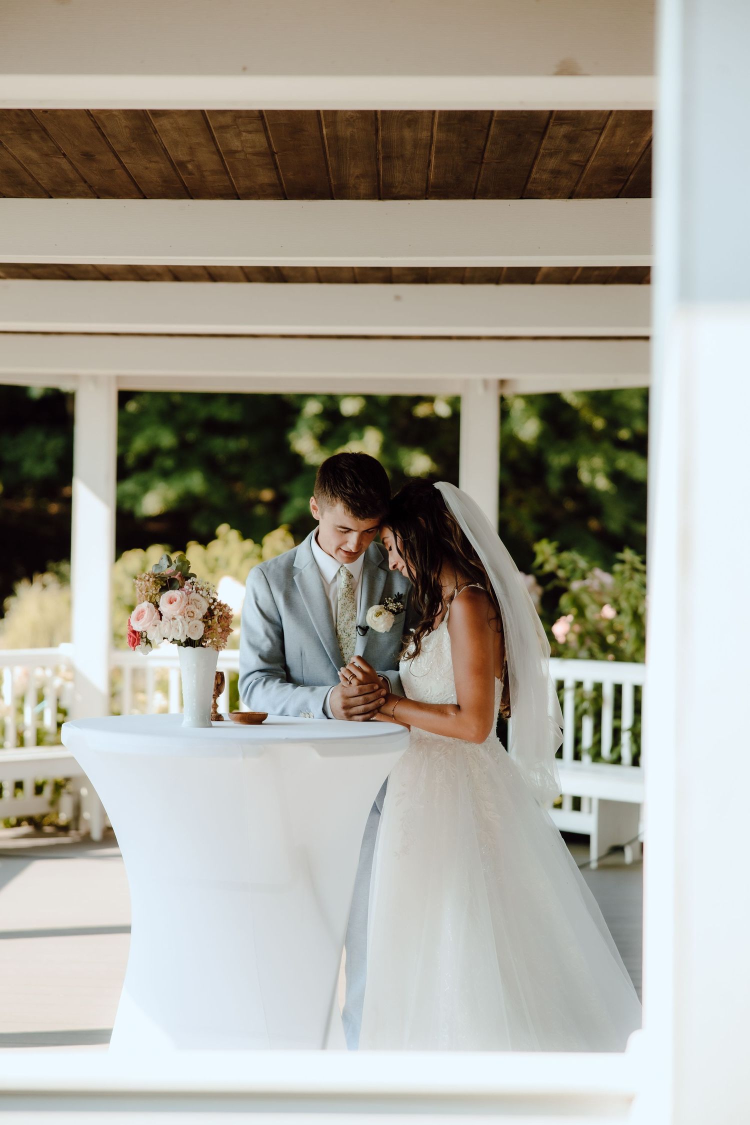 Bride and groom sharing intimate moment at cocktail table inside elegant white gazebo.