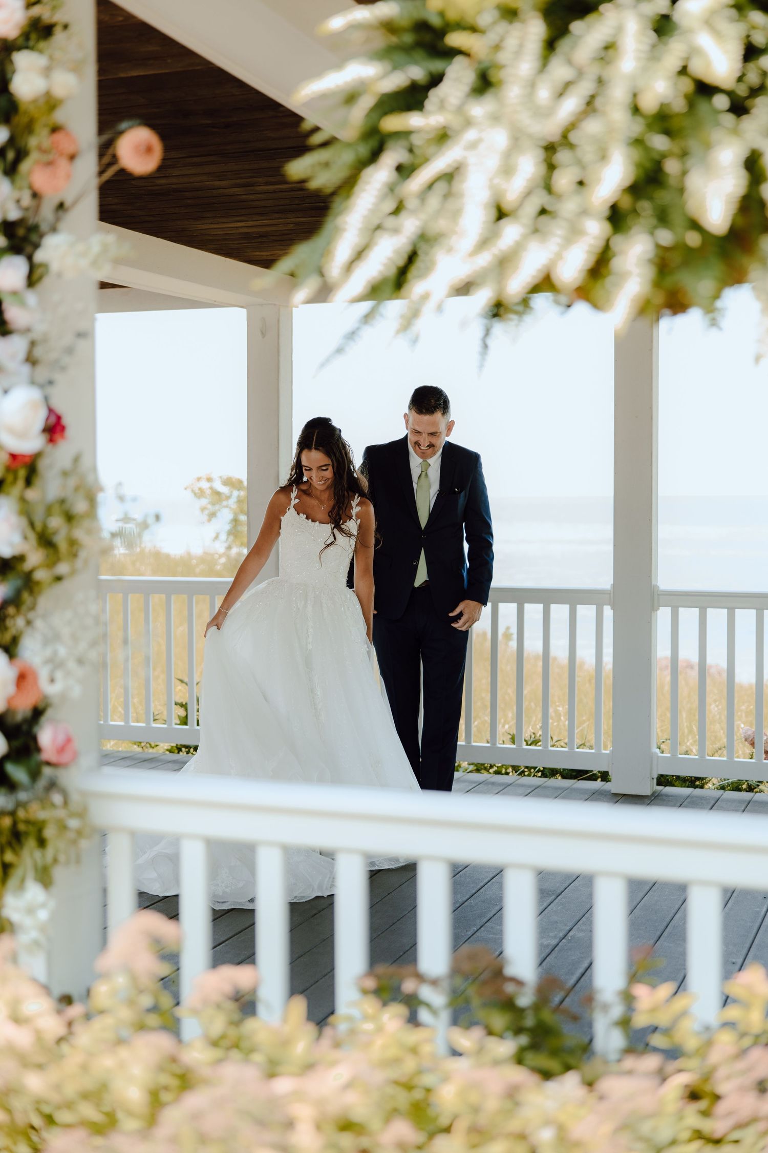 Bride and groom walk hand in hand on a decorated porch overlooking scenic water views.