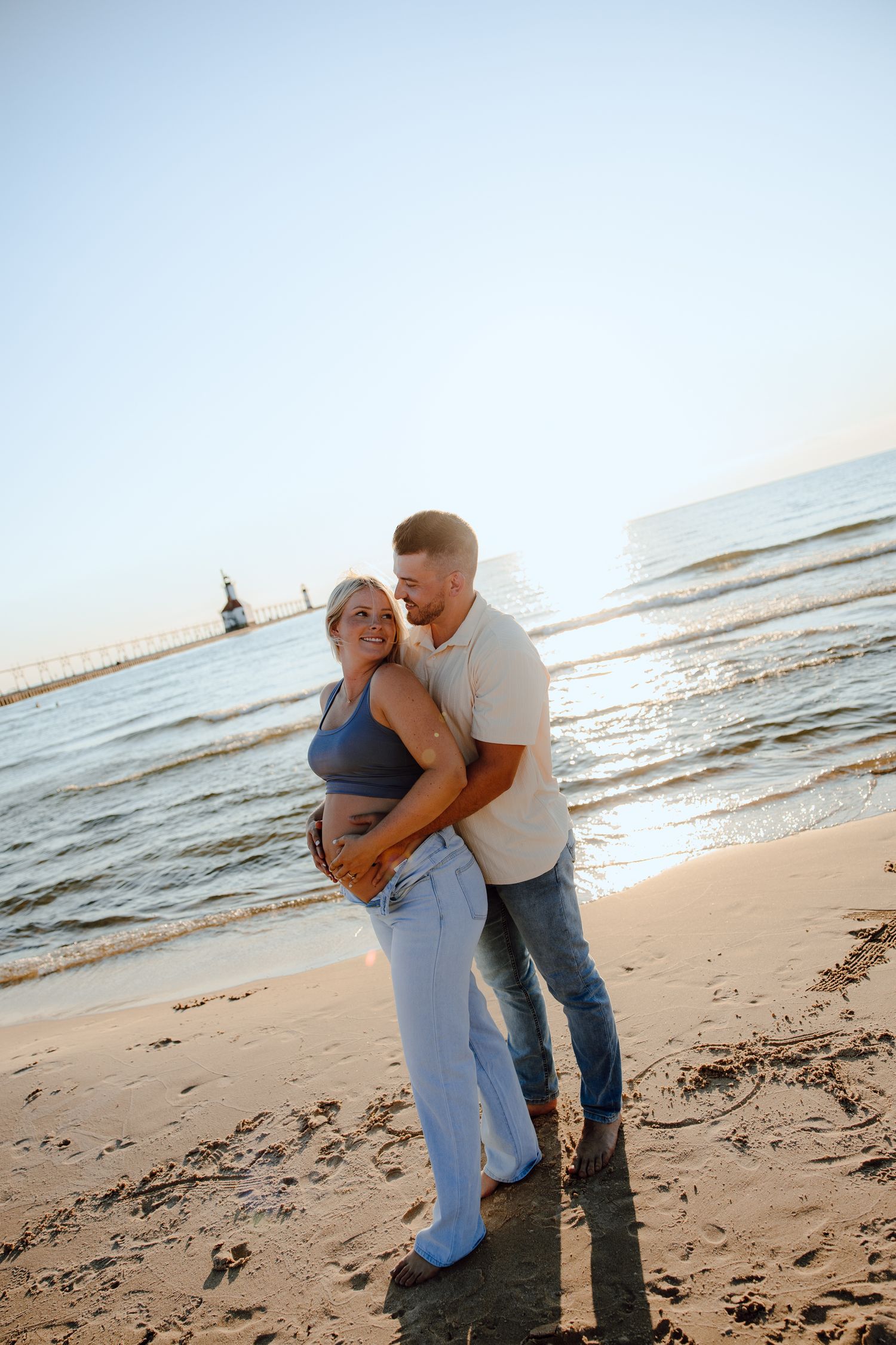Couple embraces on beach at sunset with waves and lighthouse in background.
