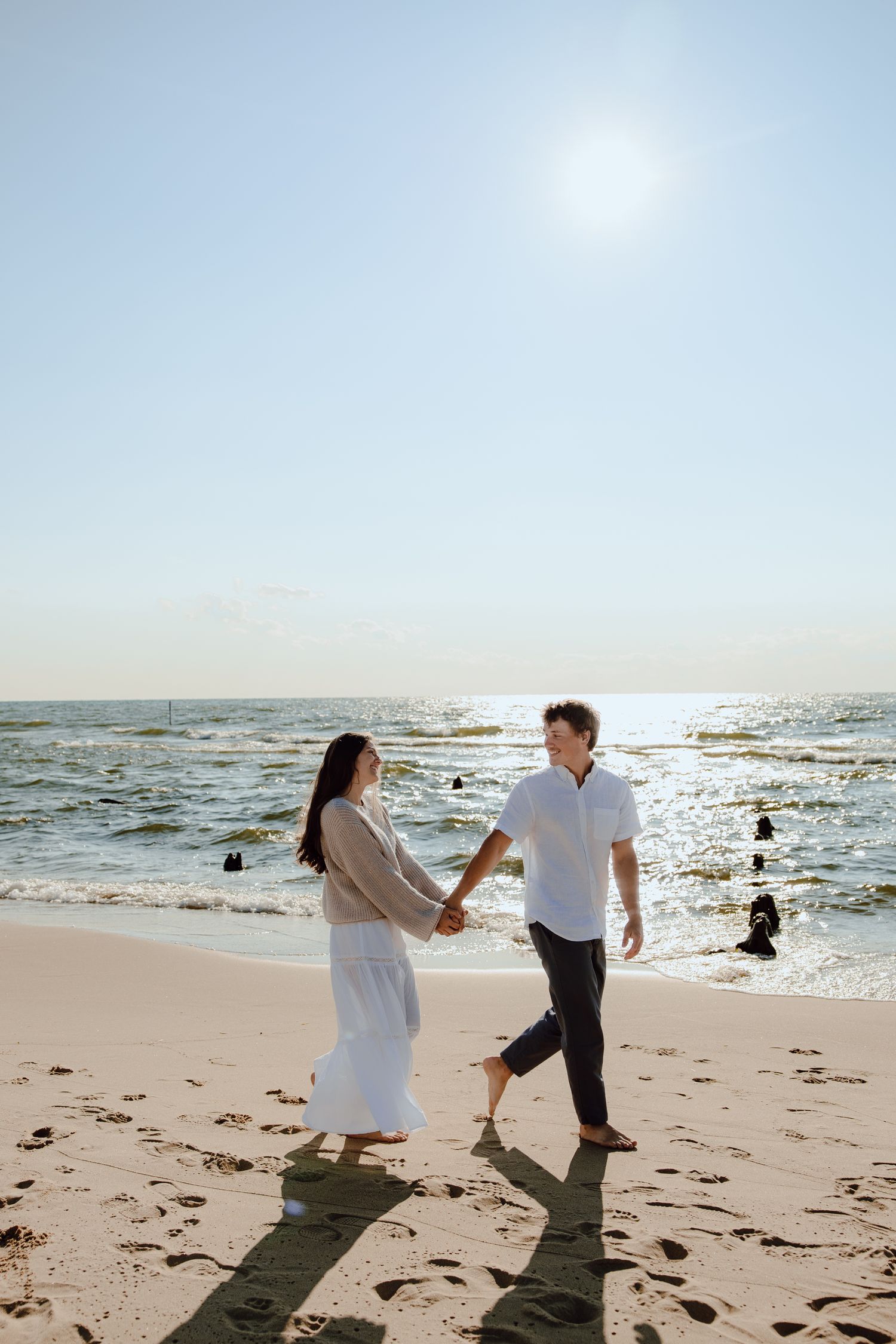 A couple holds hands while walking along a sunny beach with sparkling ocean waves.