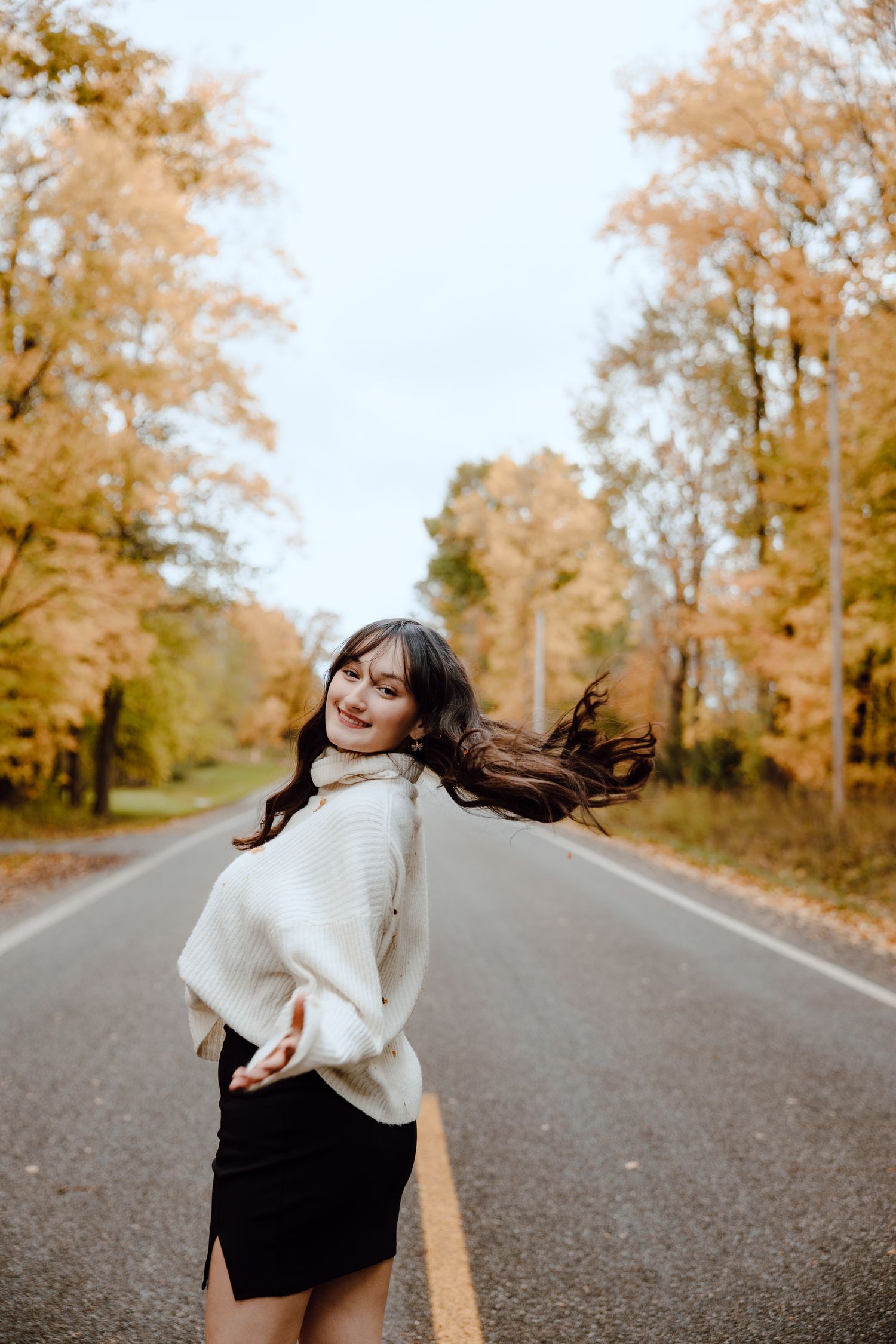 Person in white sweater twirls on empty road lined with fall trees.