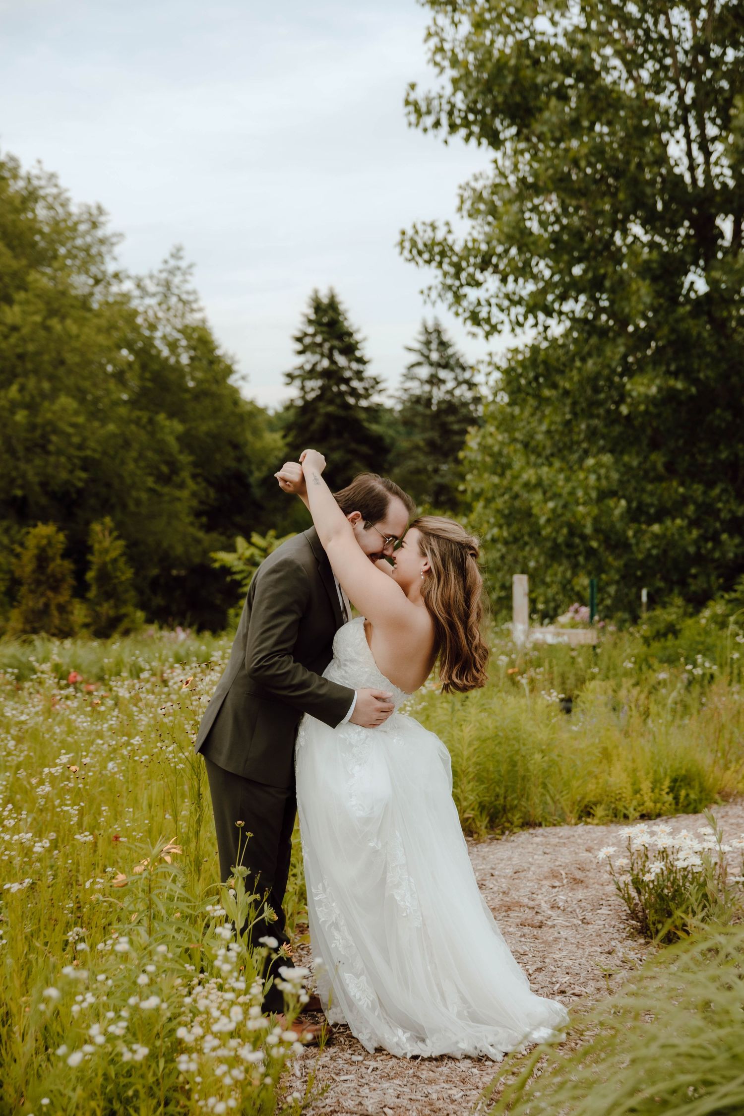 Bride and groom share a romantic kiss in a wildflower garden, with lush green trees in the background.