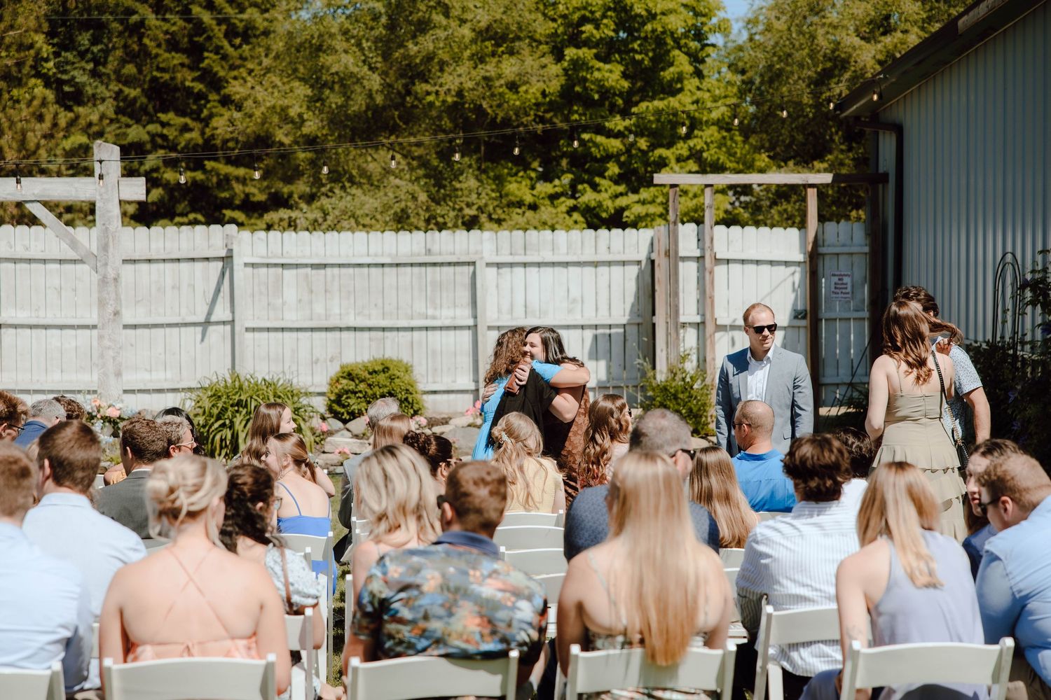 Outdoor wedding ceremony with guests seated in white chairs facing a couple embracing near a wooden fence.