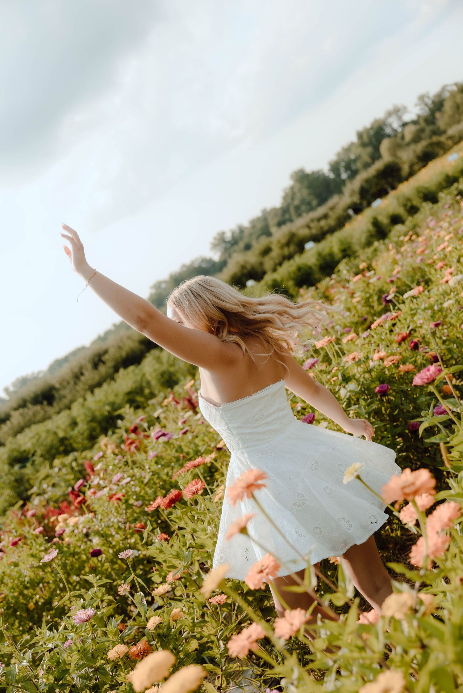 Blonde woman in white dress dancing freely in a vibrant flower field under a cloudy sky.