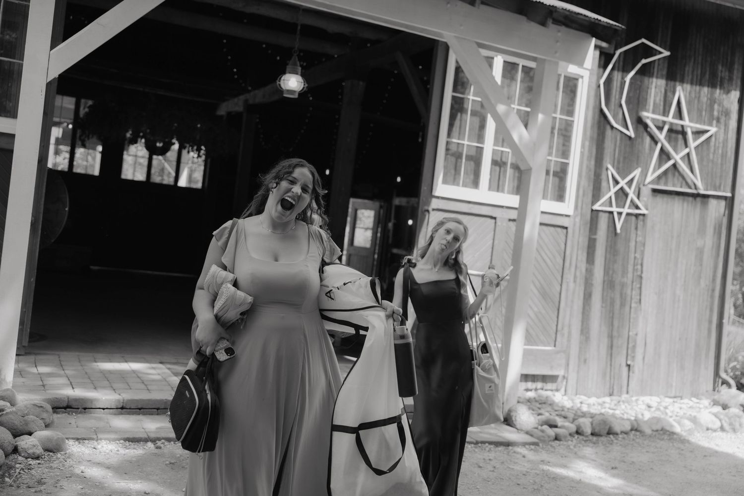 Two women in elegant dresses exit a rustic barn decorated with star and moon ornaments in black and white photo.