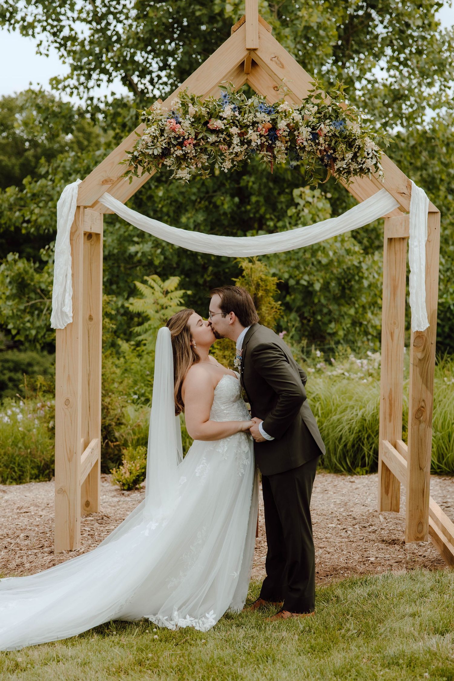 Bride and groom share first kiss under a wooden arch adorned with wildflowers and white draping.