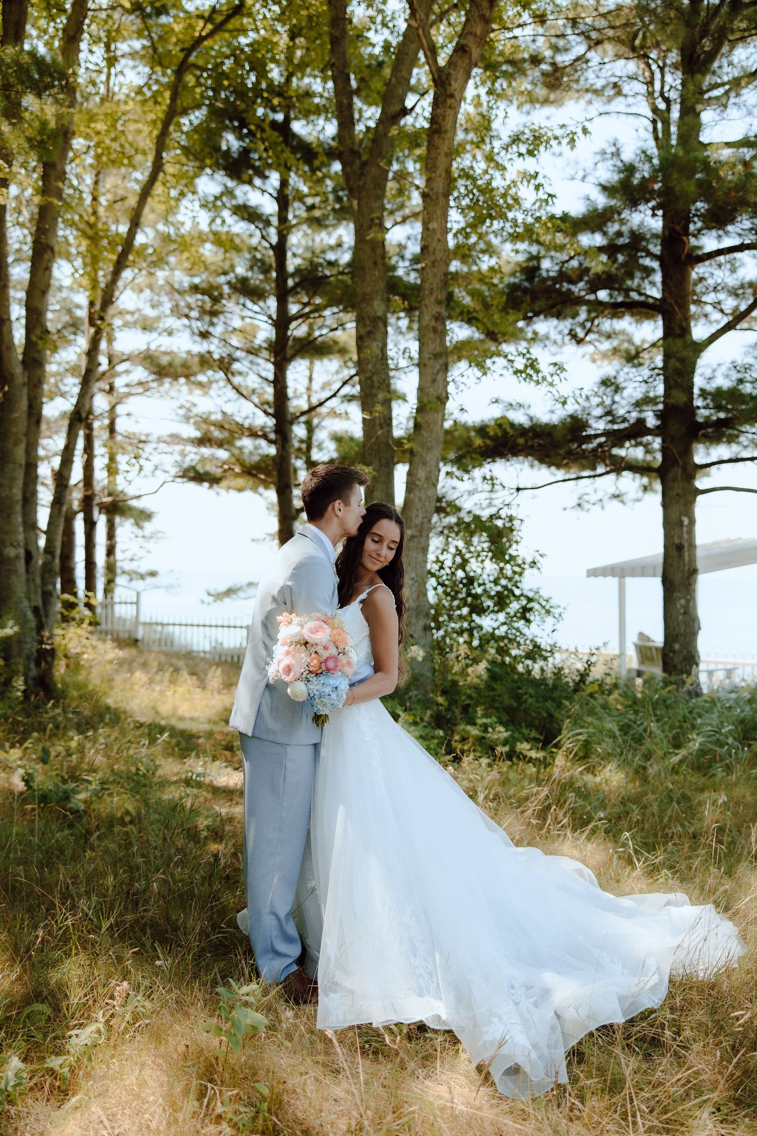Bride and groom embrace in a sunlit forest clearing, her white gown trailing on golden grass, holding a pink bouquet.