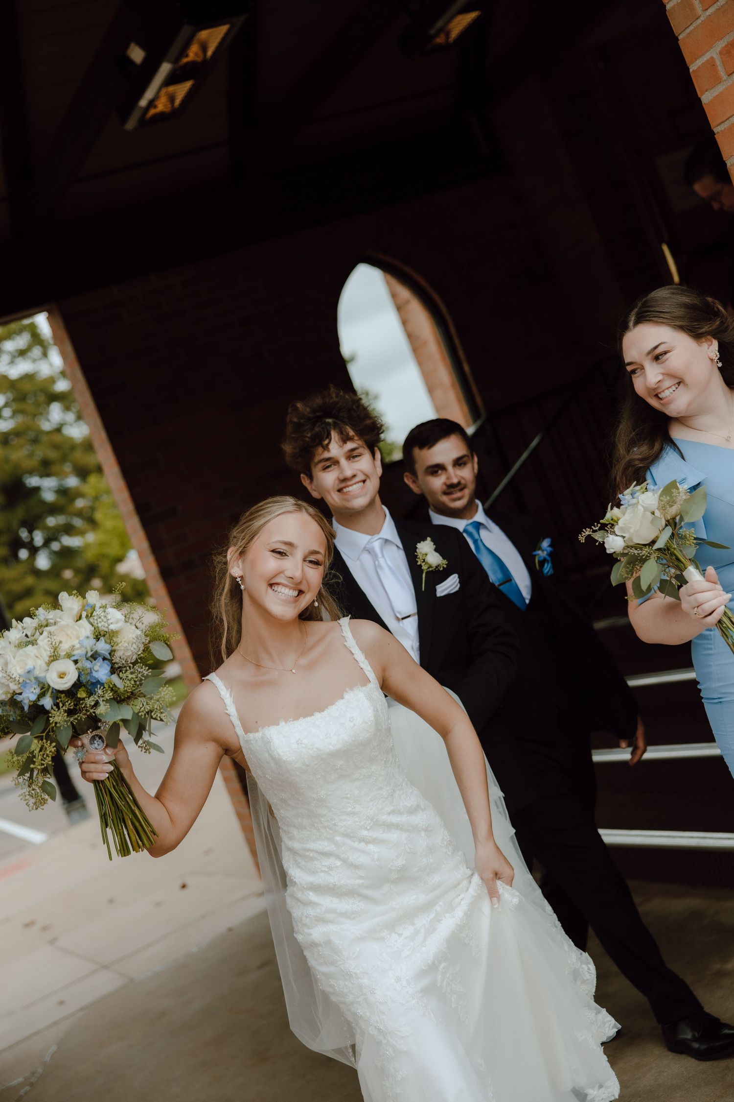 Smiling bride in white gown holds bouquet with groom and wedding party on church steps.