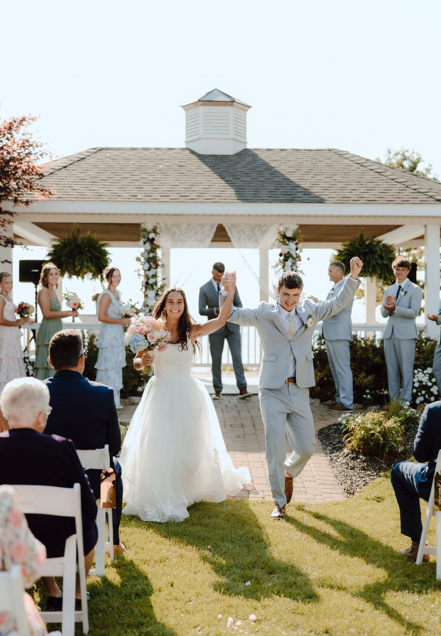 Joyful newlyweds celebrate their outdoor wedding ceremony exit near a white gazebo with bridesmaids and groomsmen cheering.