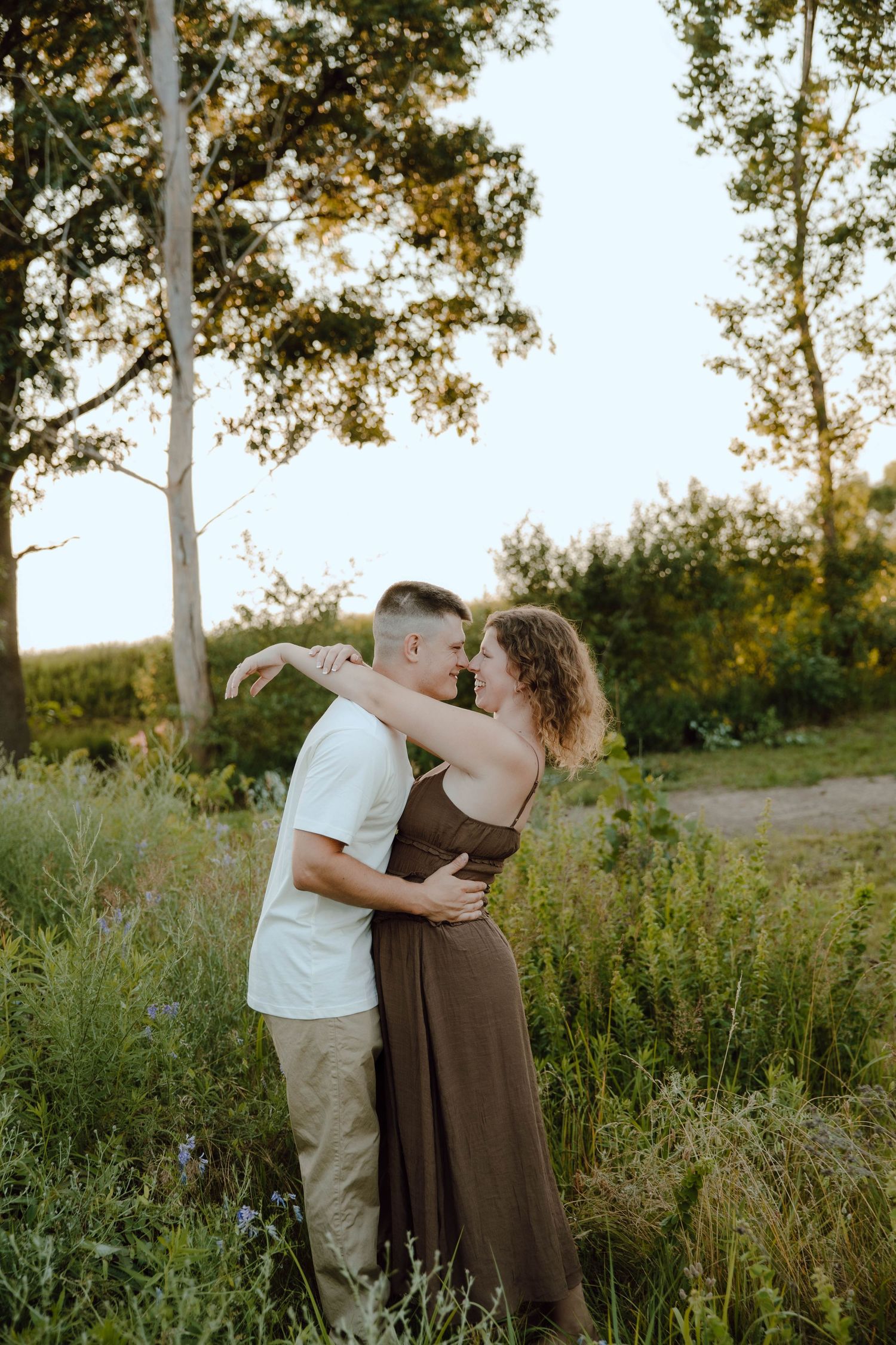 A couple embraces and shares a romantic moment in a lush green field surrounded by tall trees at golden hour.