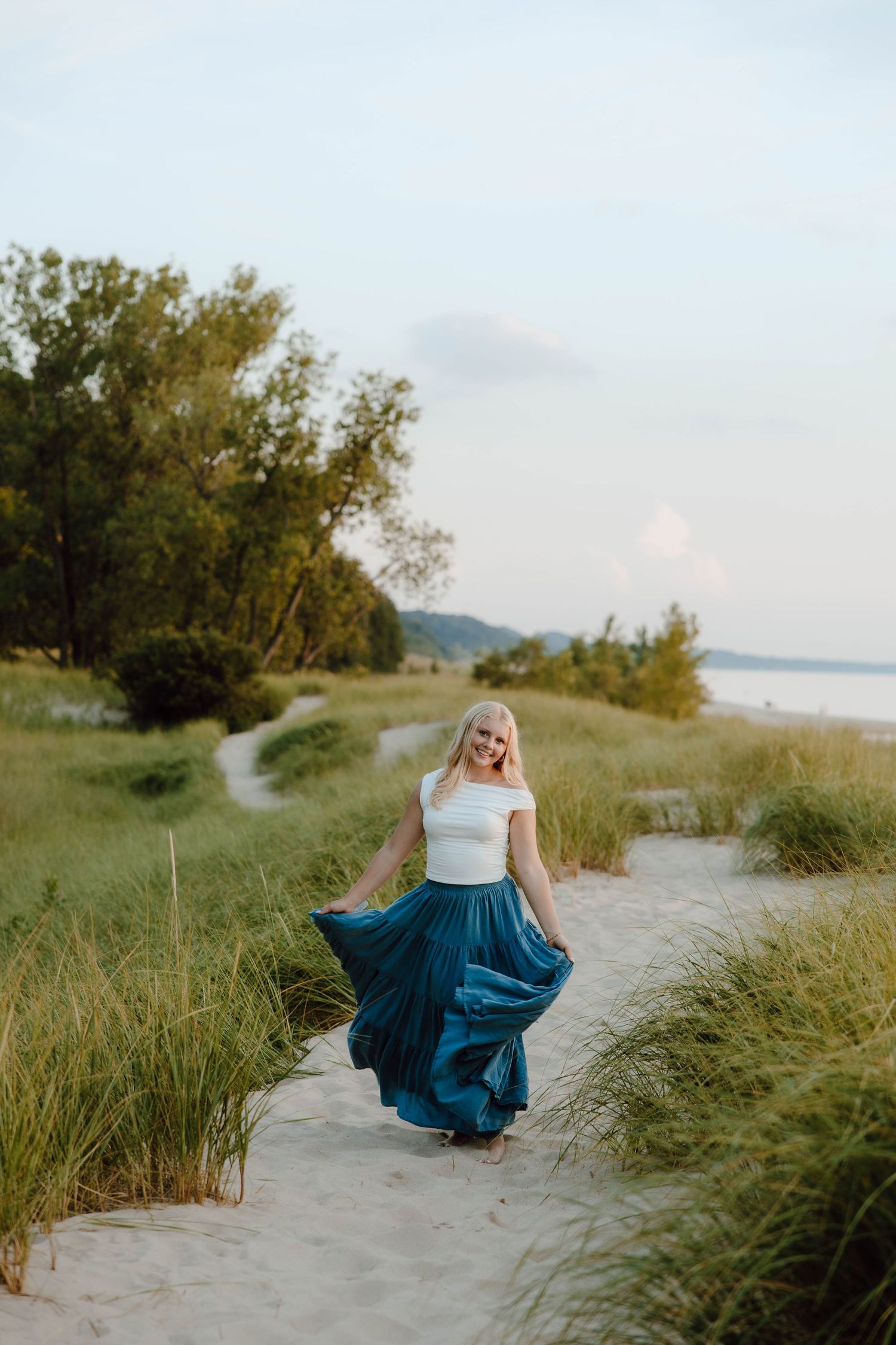 Blonde woman twirling in a teal maxi skirt on a sandy beach path surrounded by green dunes and trees.