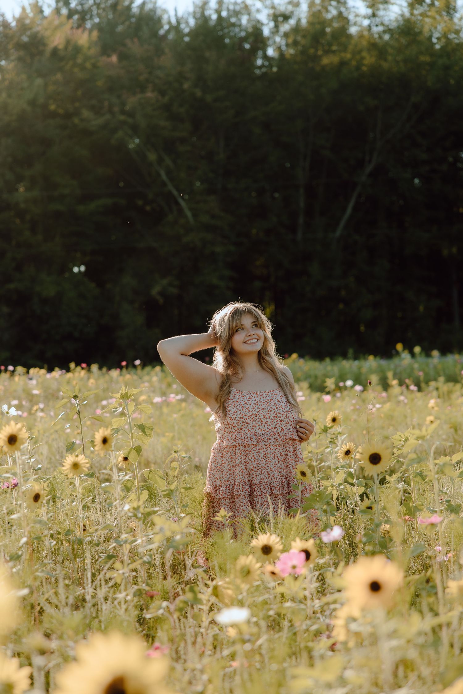 Young blonde girl in floral dress smiling in sunlit wildflower field with sunflowers and trees.