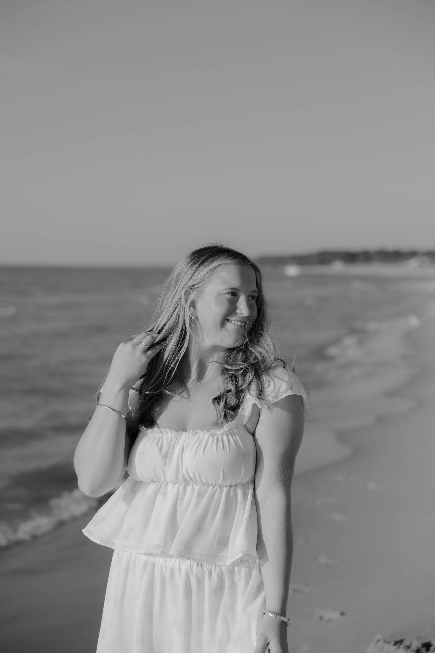 Black and white photo of a smiling woman in a white dress standing on a beach with ocean waves behind her.