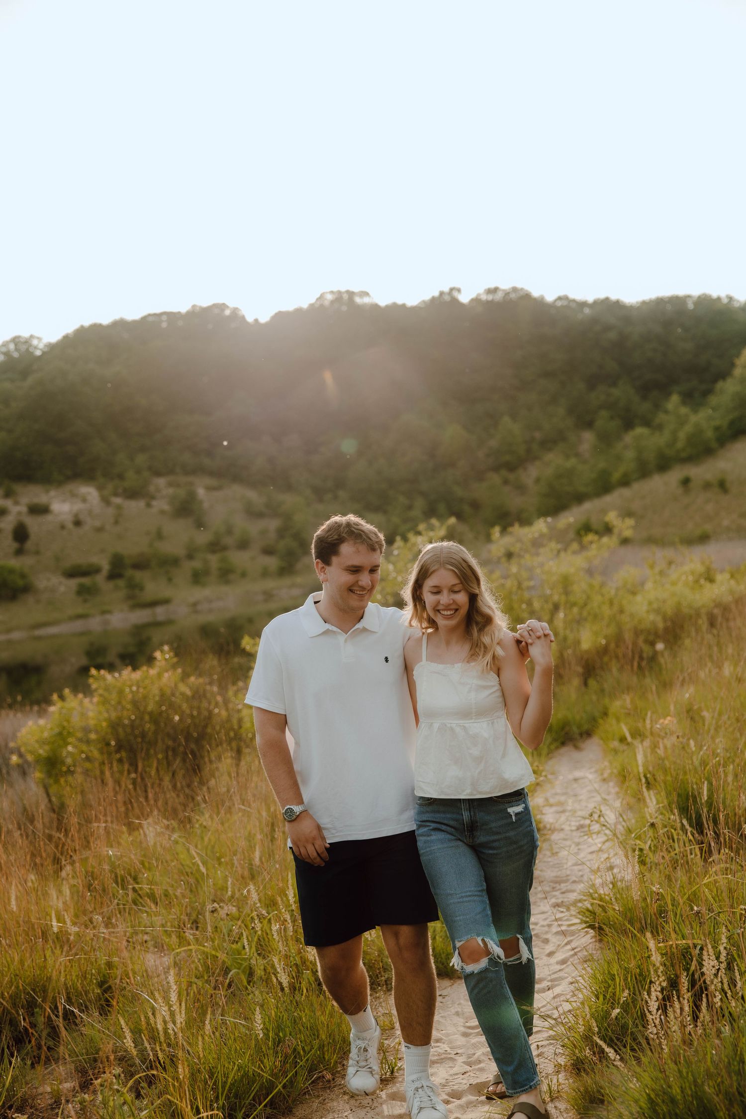 A smiling couple walks hand in hand on a sunlit trail surrounded by lush green hills and wildflowers.