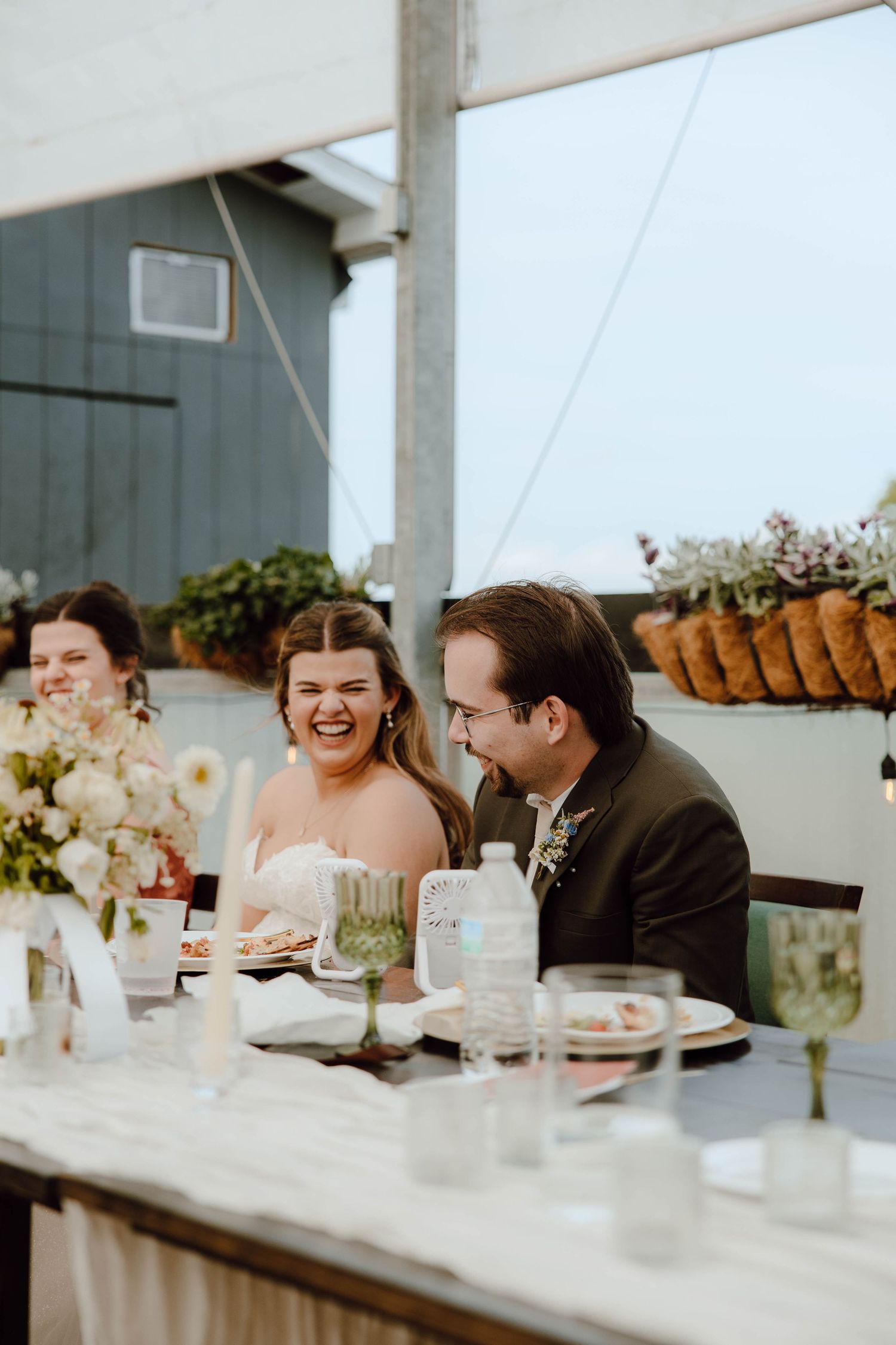 Bride and groom laugh joyfully at sweetheart table during outdoor wedding reception.
