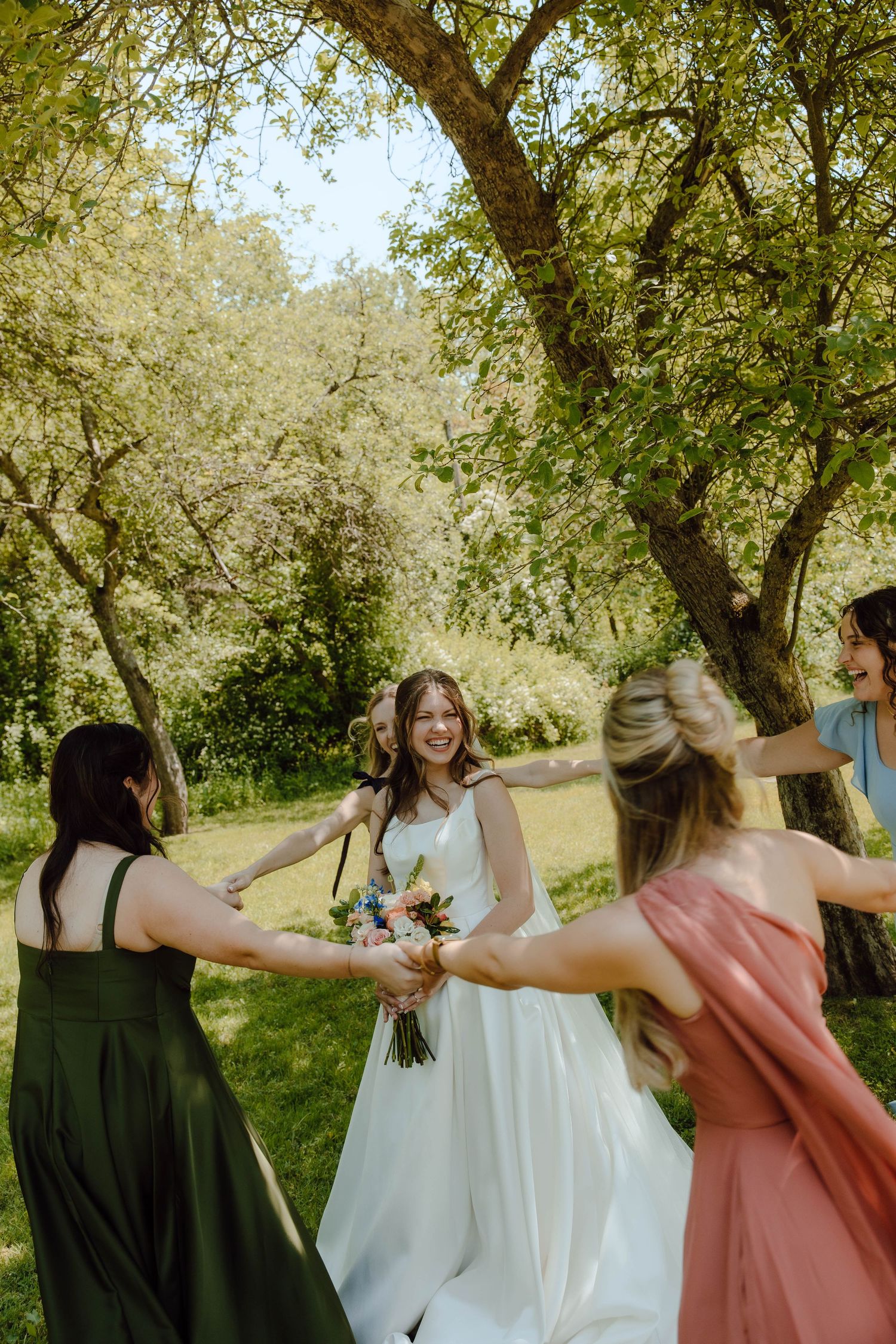 Bride in white gown laughs as bridesmaids in colorful dresses hold hands dancing together outdoors.