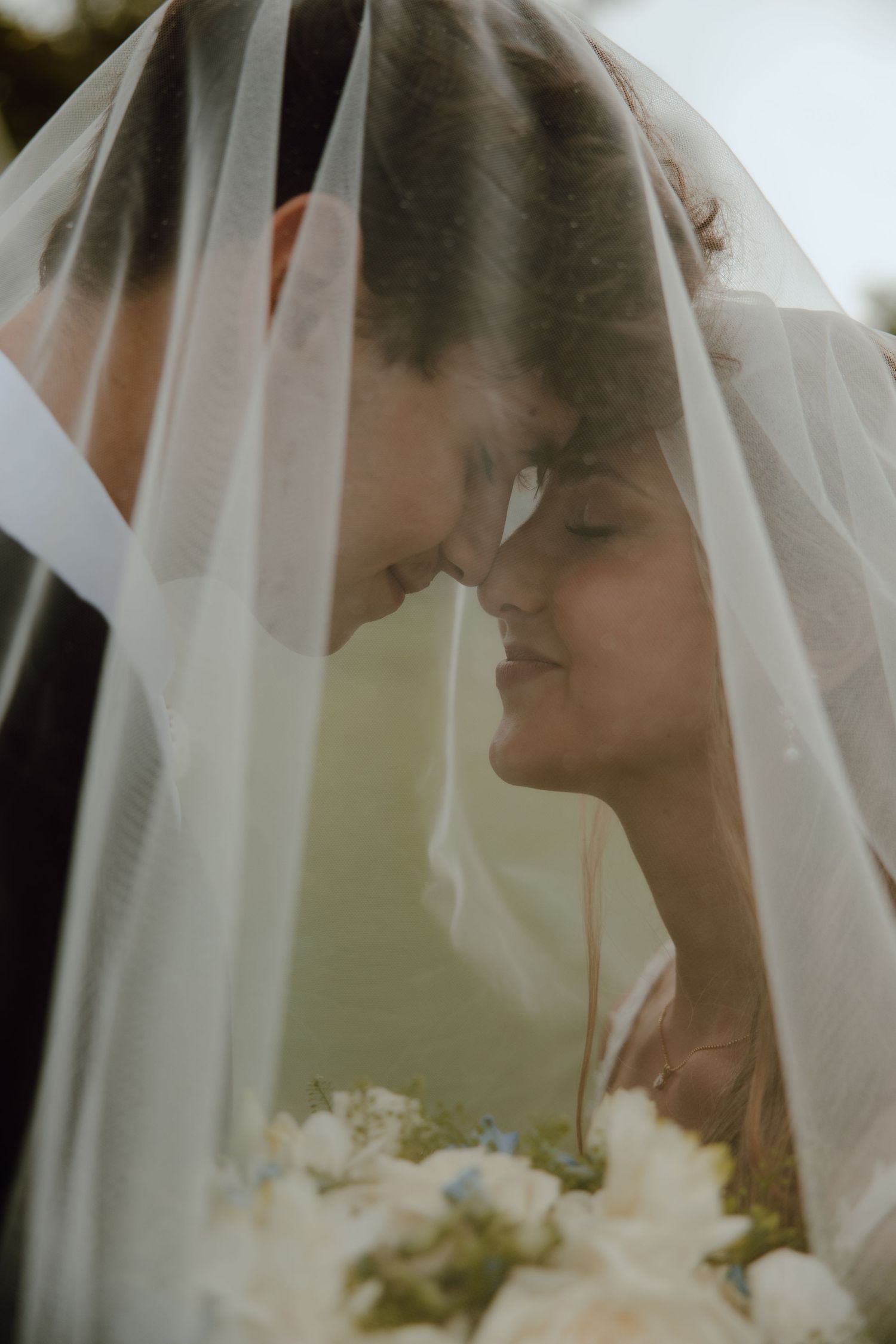 Romantic bride and groom moment under wedding veil with bouquet.