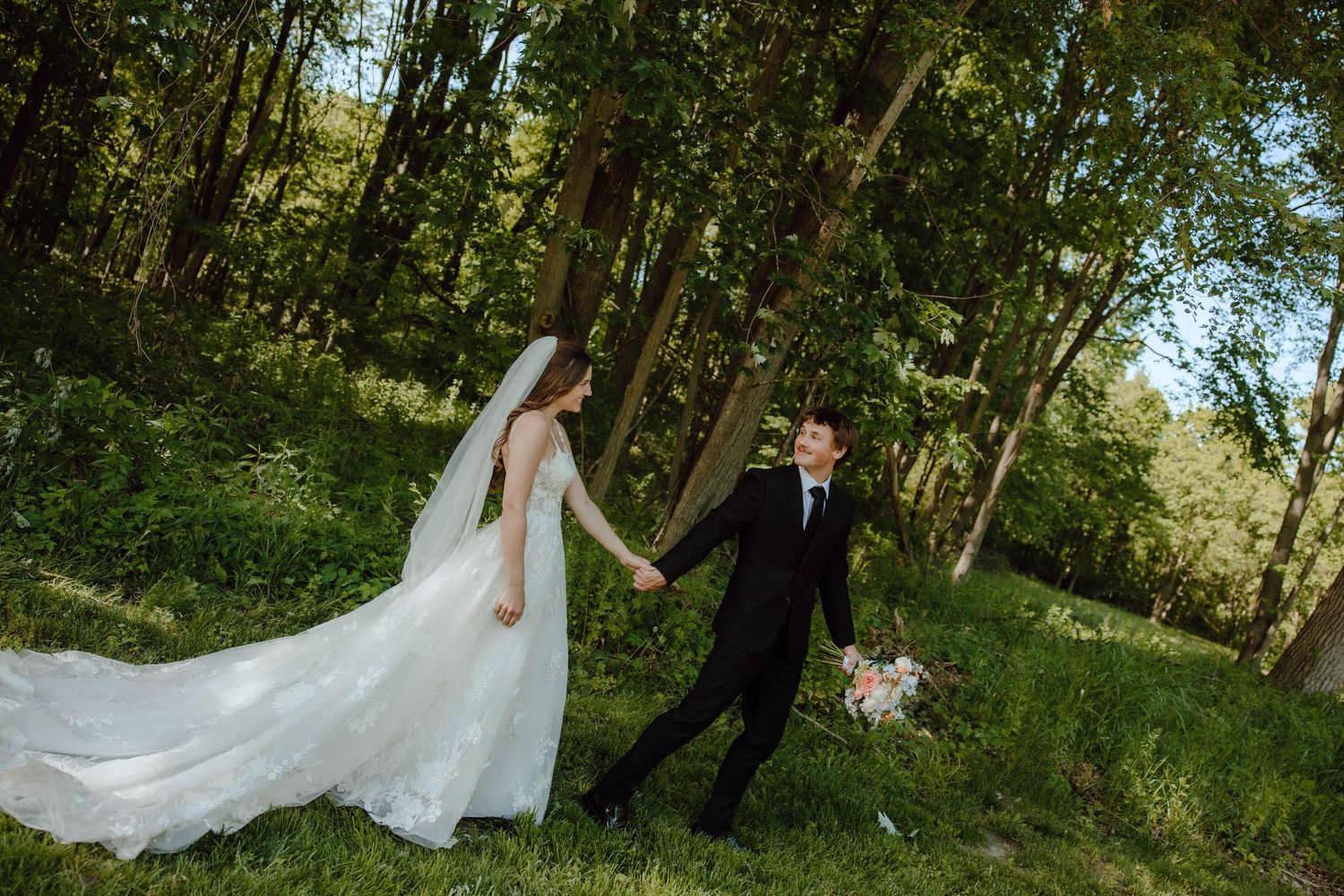 Bride and groom walk hand-in-hand through a lush green forest, bride's white gown trailing behind her.