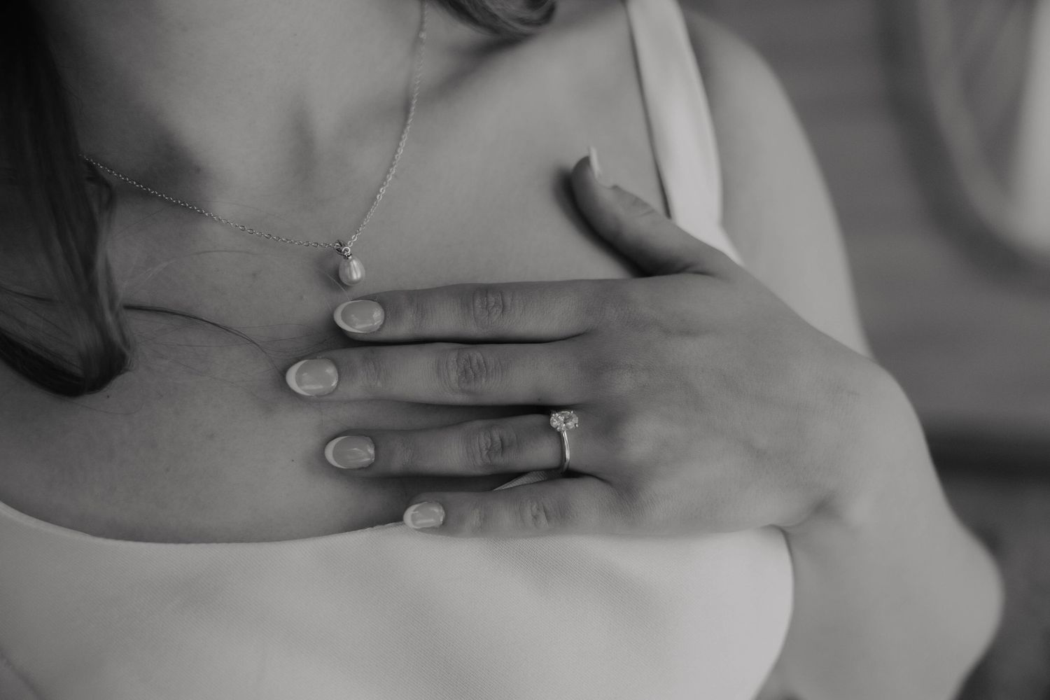 Black and white close-up of a woman's hand with a diamond ring resting on her chest near a delicate necklace.