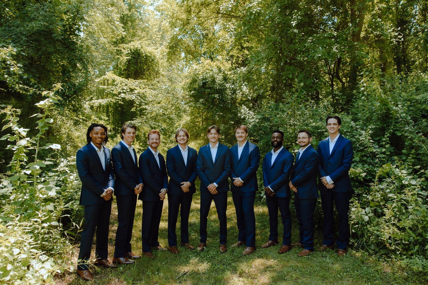 Group of groomsmen in navy suits posing outdoors among lush green trees for a wedding photo.