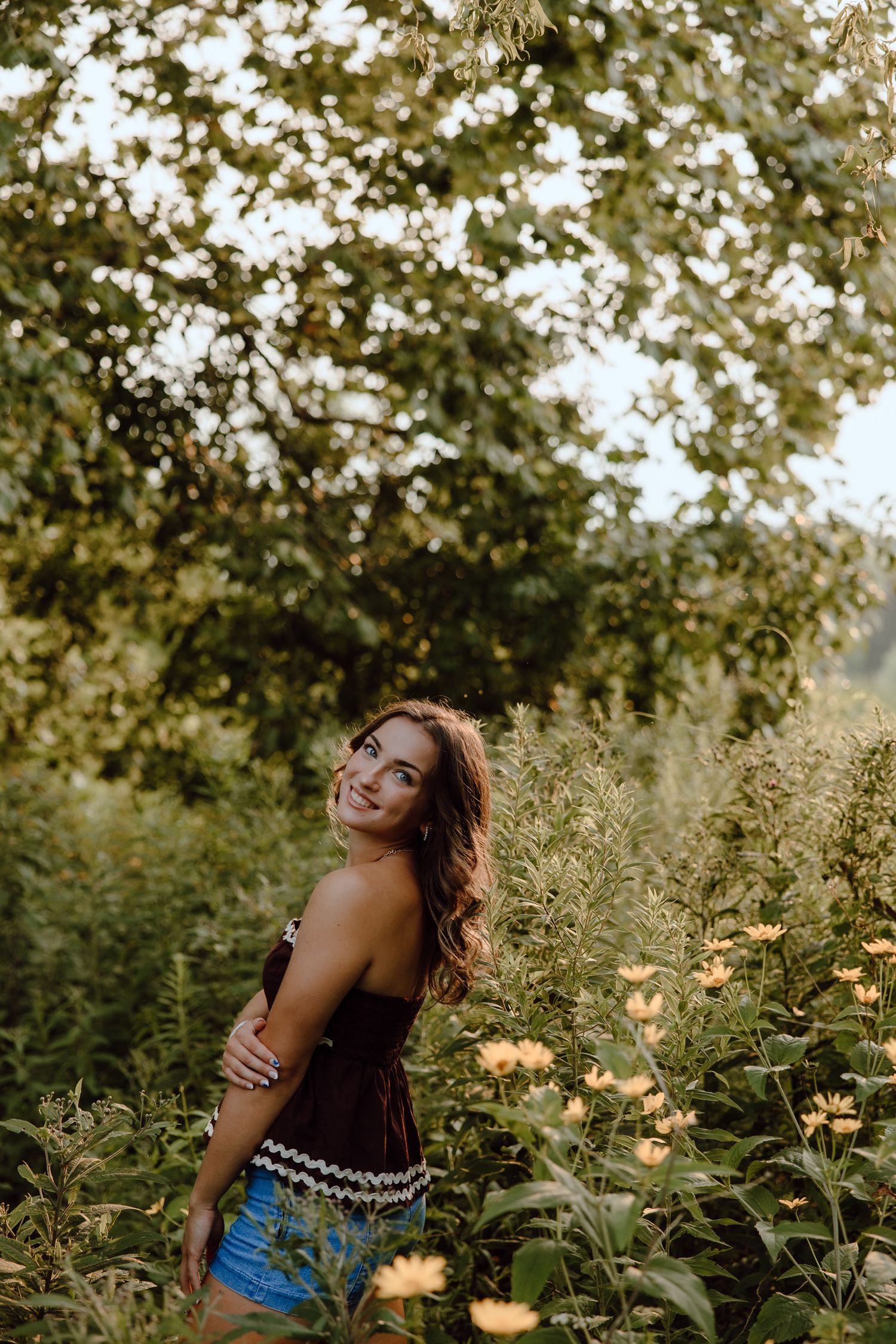 A person in a black top and denim poses joyfully in a garden with golden flowers.