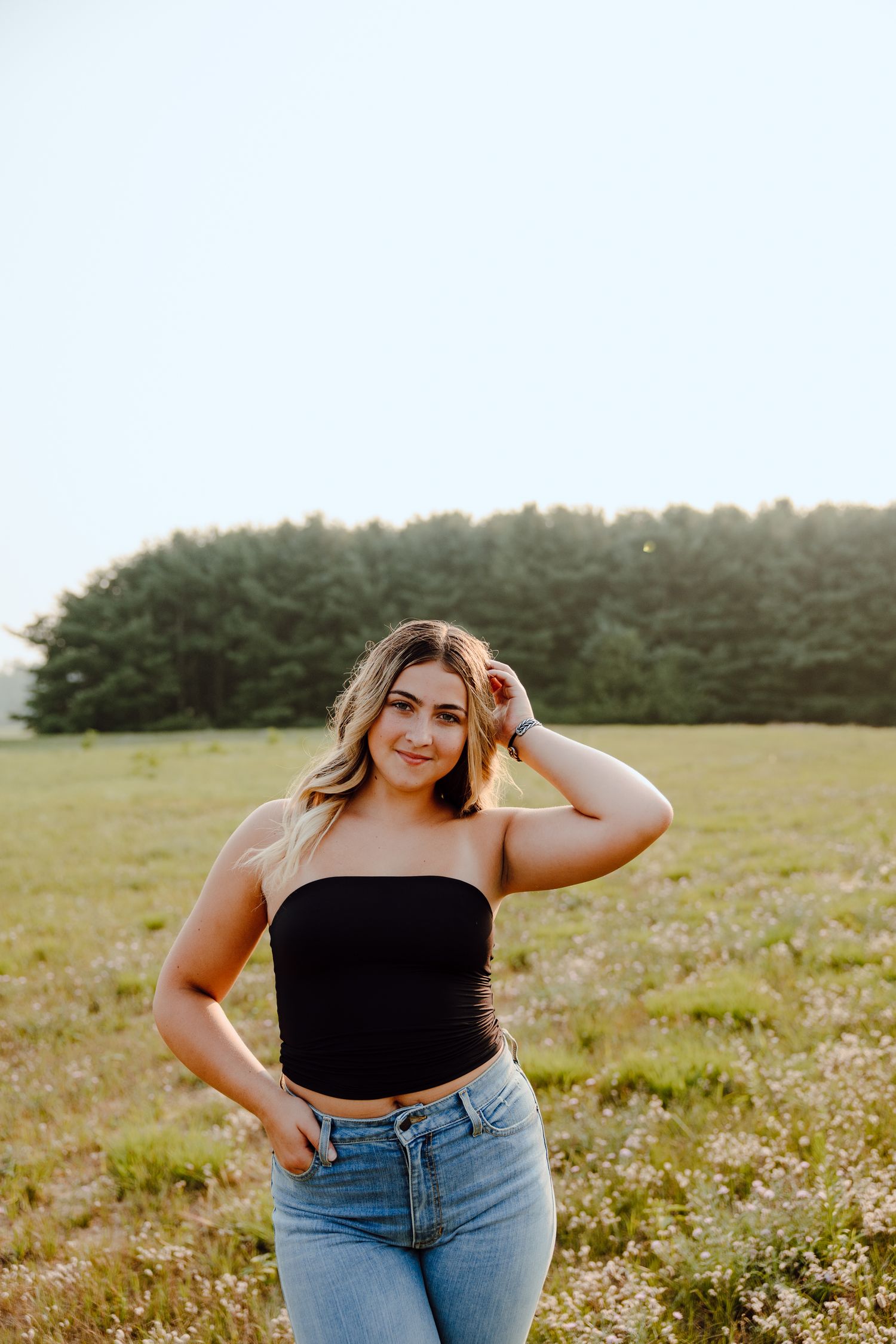Woman in black top and jeans poses confidently in a wildflower meadow at sunset.