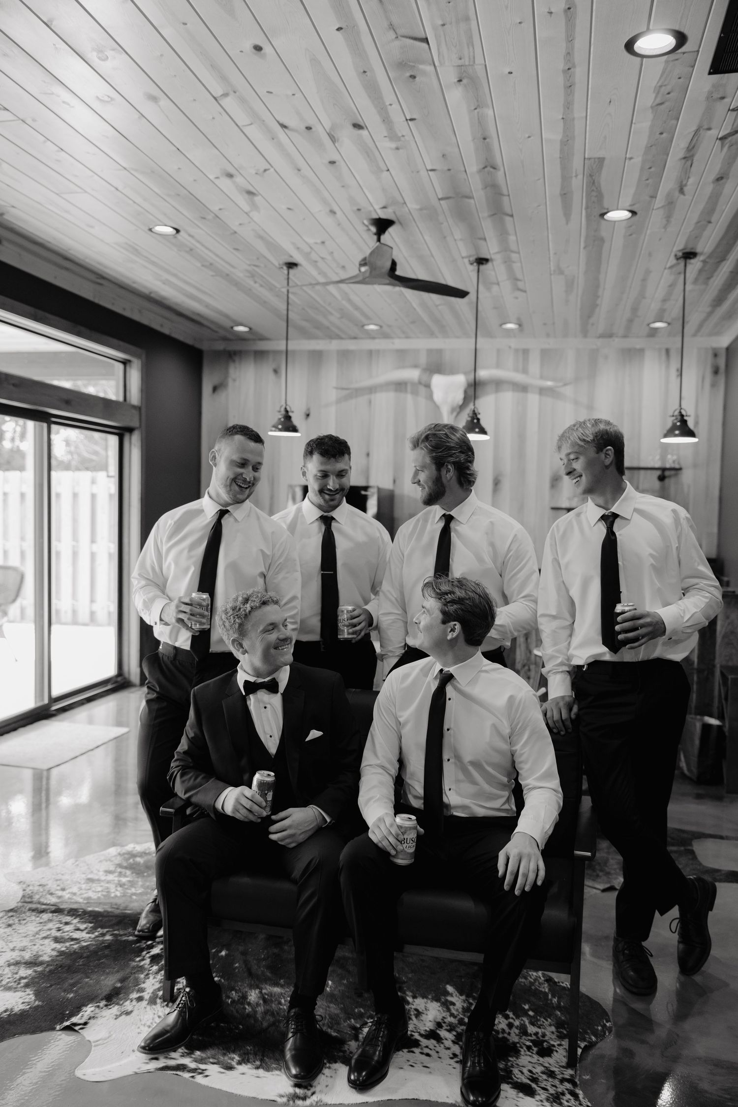 Groom and groomsmen laughing together in black and white photo, dressed in suits and ties before wedding ceremony.