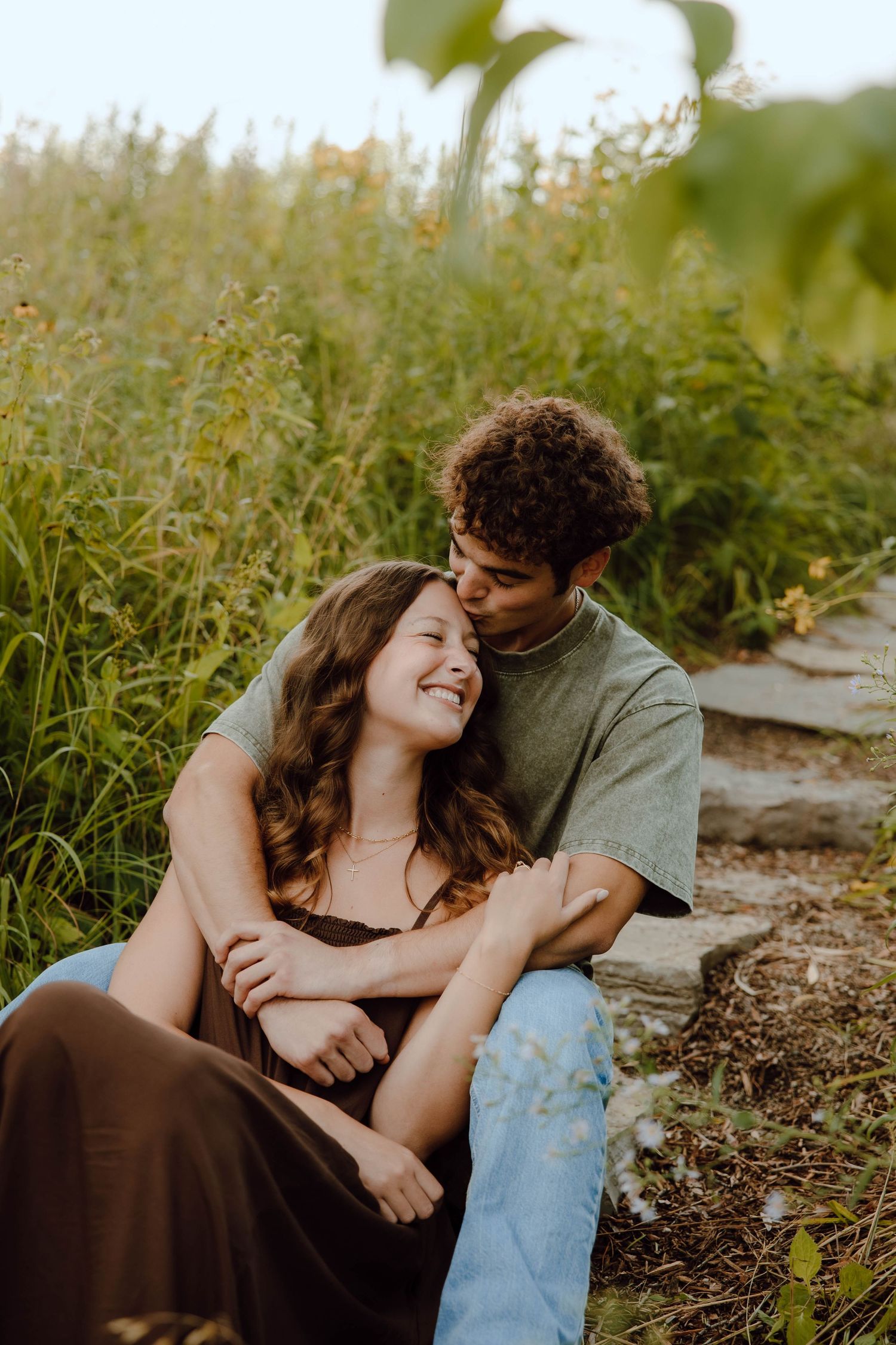 A couple sits together in a wildflower field, the man embracing the woman as she smiles up at him.
