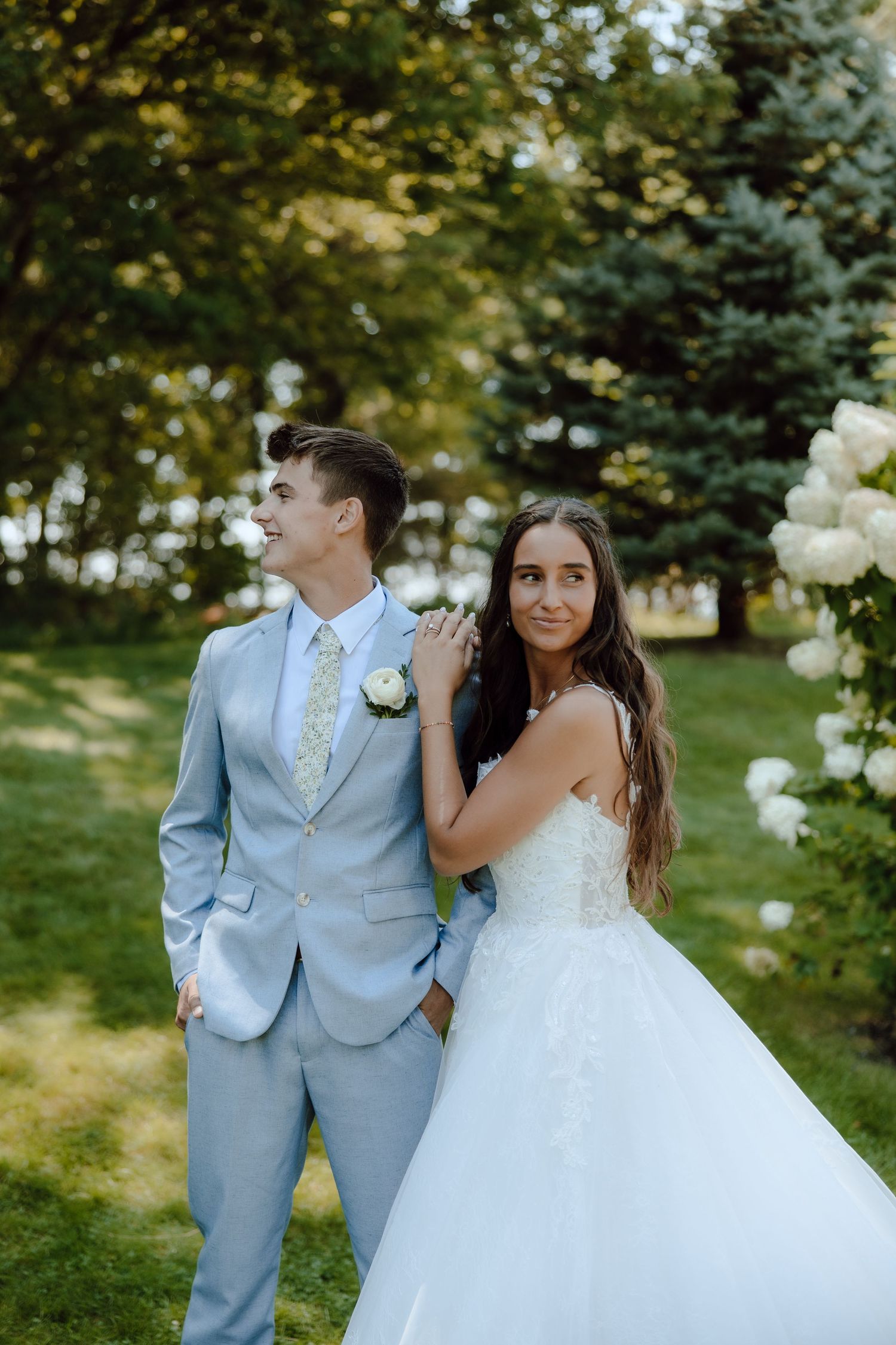 Bride and groom pose outdoors in lush garden, groom in light blue suit, bride in white gown.