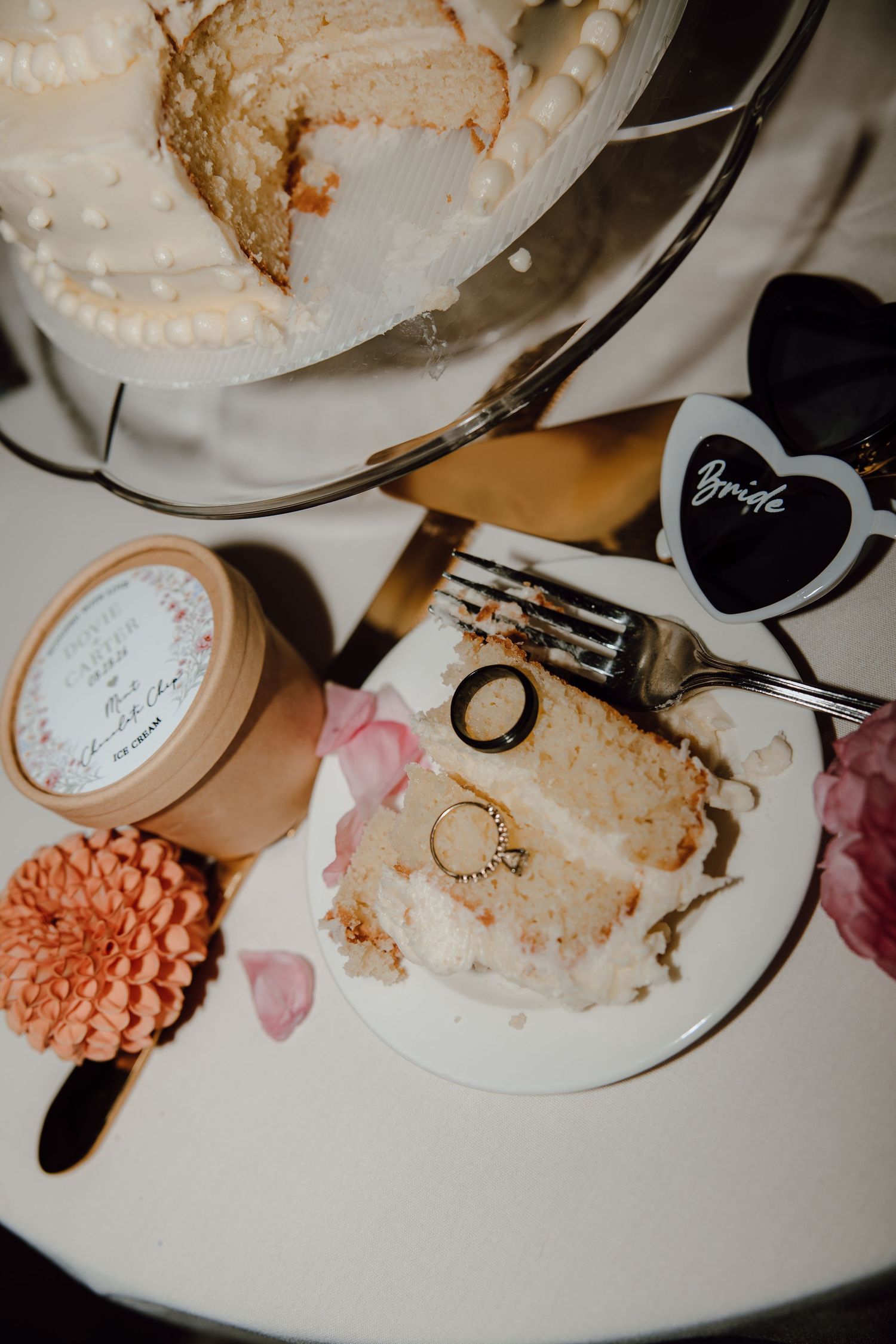 Wedding cake slice on plate with bride label, fork, rings, and floral decor on white table.