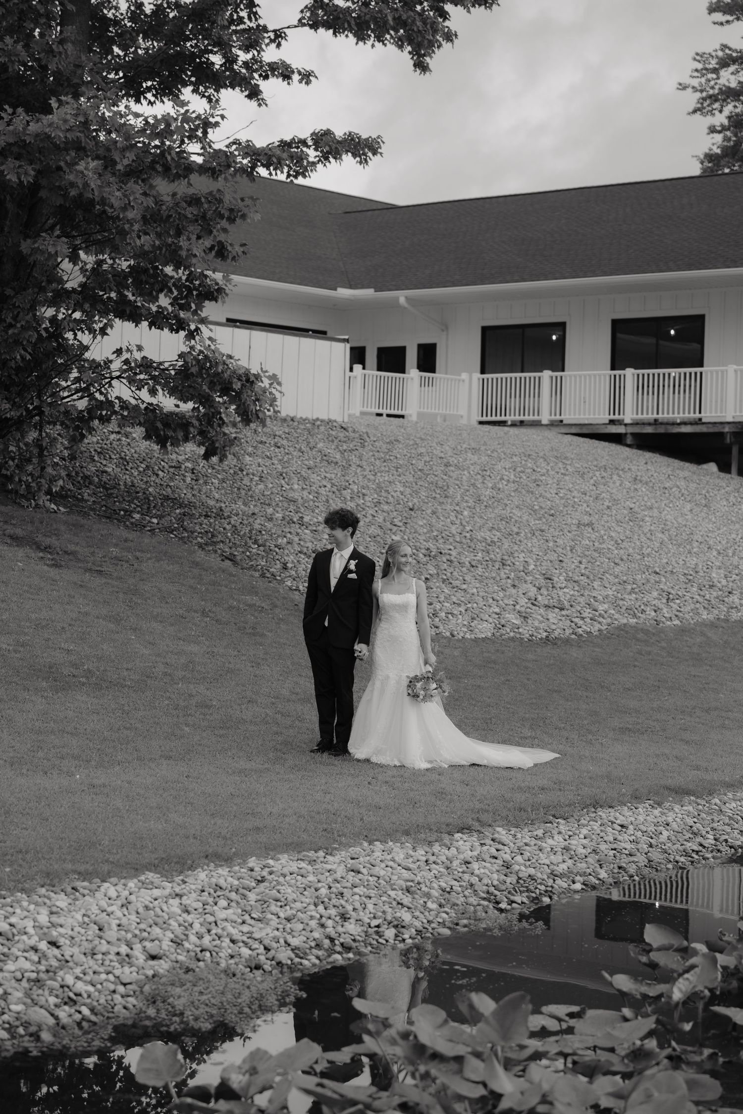 Black and white photo of bride and groom standing by pond near wedding venue.