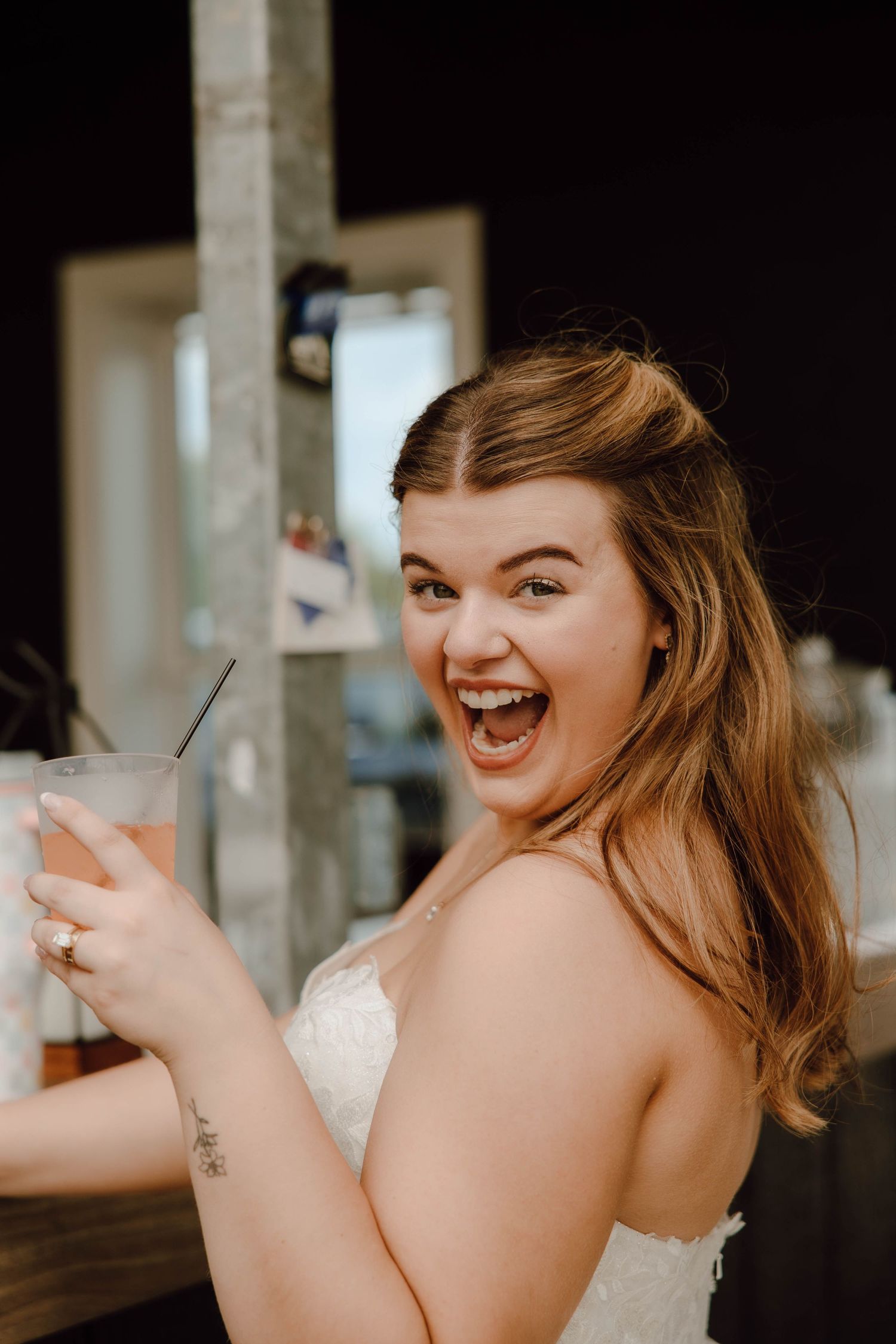 Excited bride in white dress laughing while holding a drink at her wedding reception.