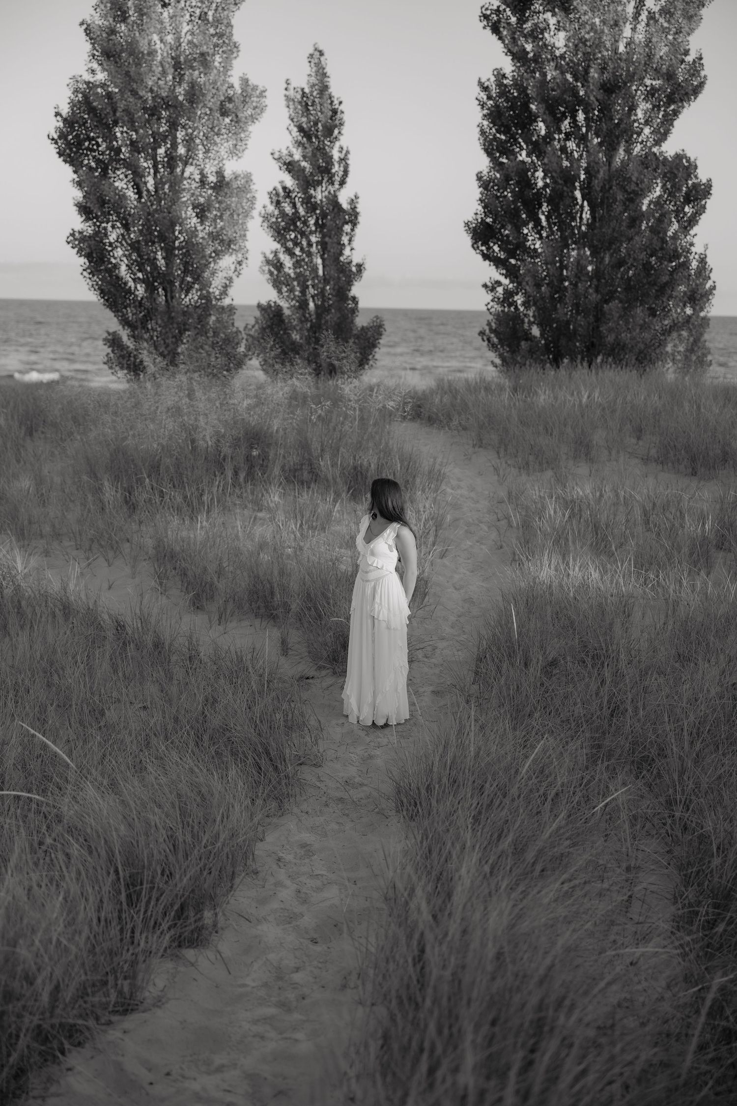 Woman in white dress walks sandy path through tall grass toward trees and ocean in black and white photo.