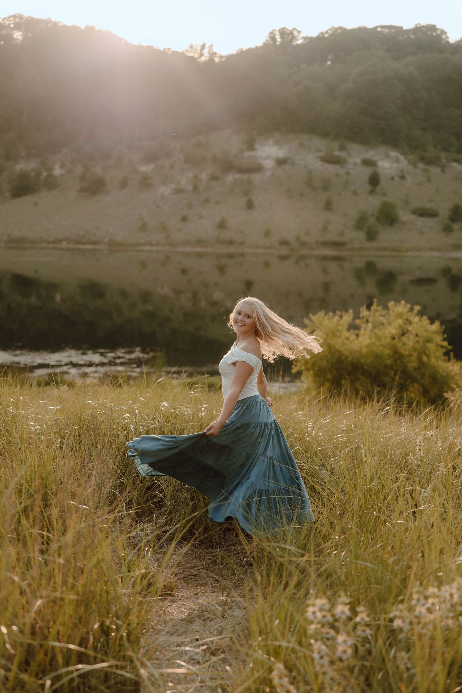 Blonde woman twirling in teal skirt and white top in golden meadow with mountain lake backdrop at sunset.